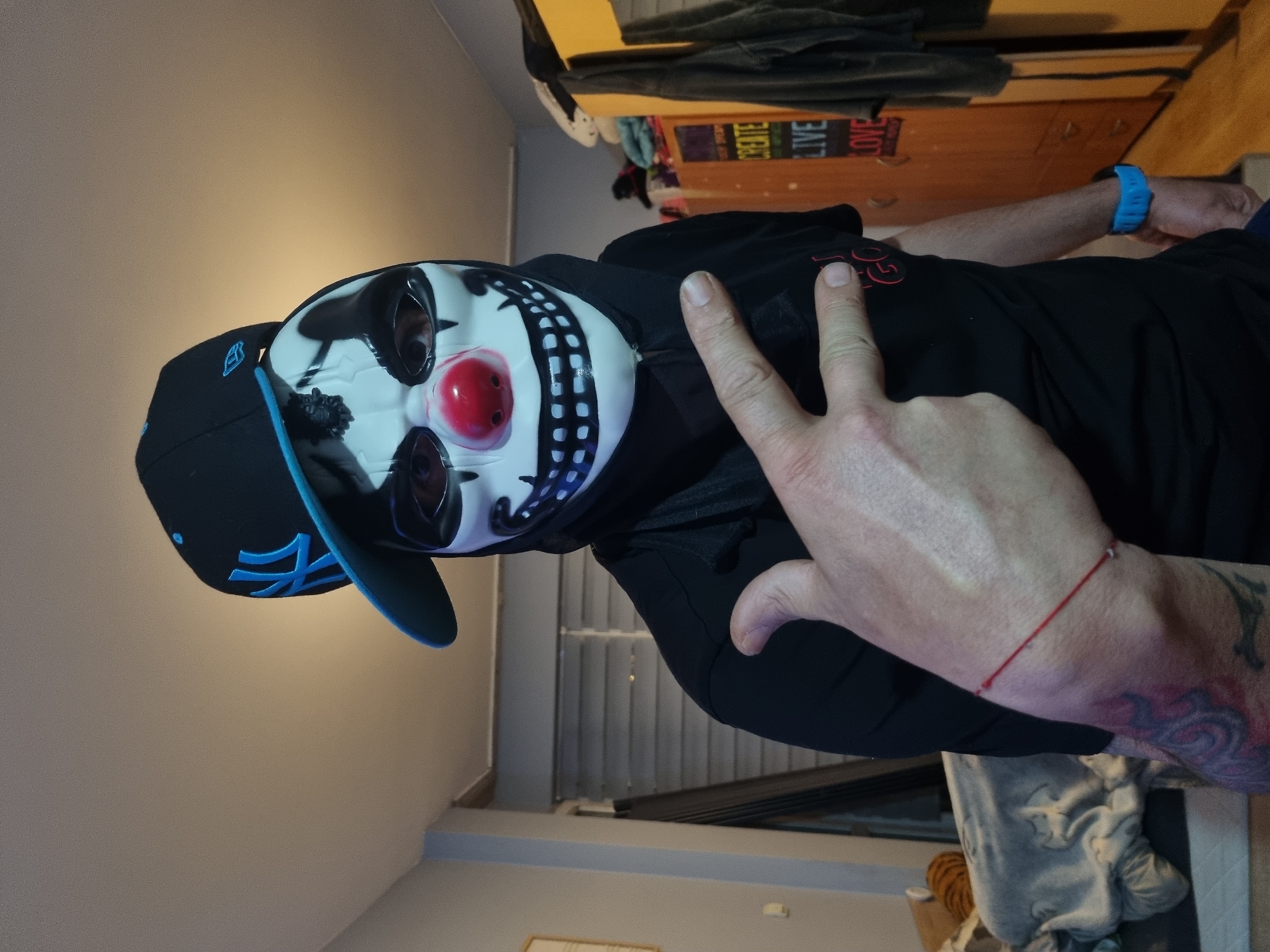 A man wearing a black baseball cap with a blue logo and a clown mask is giving a peace sign. He is also wearing a black shirt. The background is a bedroom.