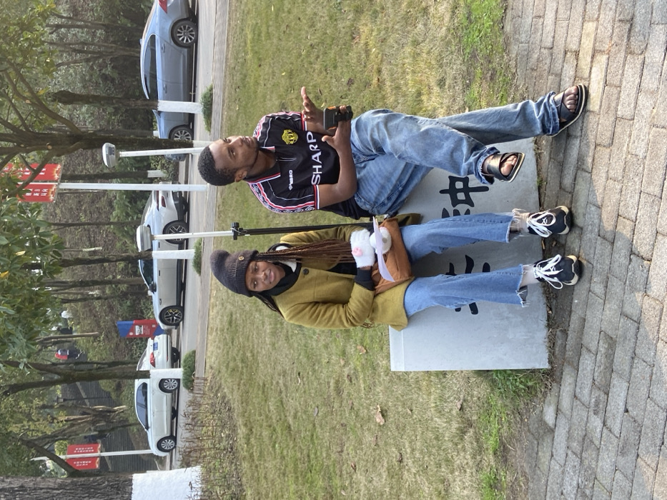 A man and woman sit on a rectangular stone object. The man is wearing jeans, sandals, and a black and red sports jersey while holding a camera. The woman wears a beanie, tan coat, blue jeans, and black and white shoes and holds a brown bag. They are outside on green grass near a brick walkway and trees with multiple cars parked nearby. The stone object they sit on has characters written on it.