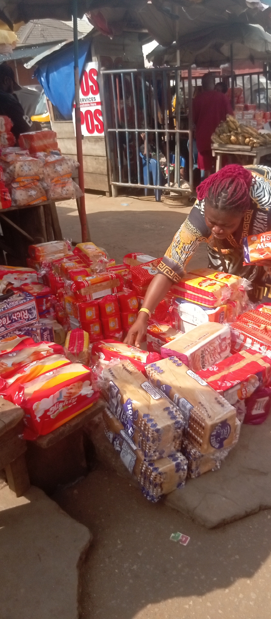 In Nkpor, Nigeria, a woman with braided burgundy hair is arranging stacks of wrapped bread loaves at an outdoor market stall. The stall is overflowing with packages of sliced bread, some labeled "Mighty" and "Rich Tea," while others display the "Hossana Magic Honey" brand. She wears a black and gold patterned top and a gold bracelet. In the background, there are other market vendors and customers, and a sign indicating "POS" services. The ground is paved and the scene is bustling with activity.