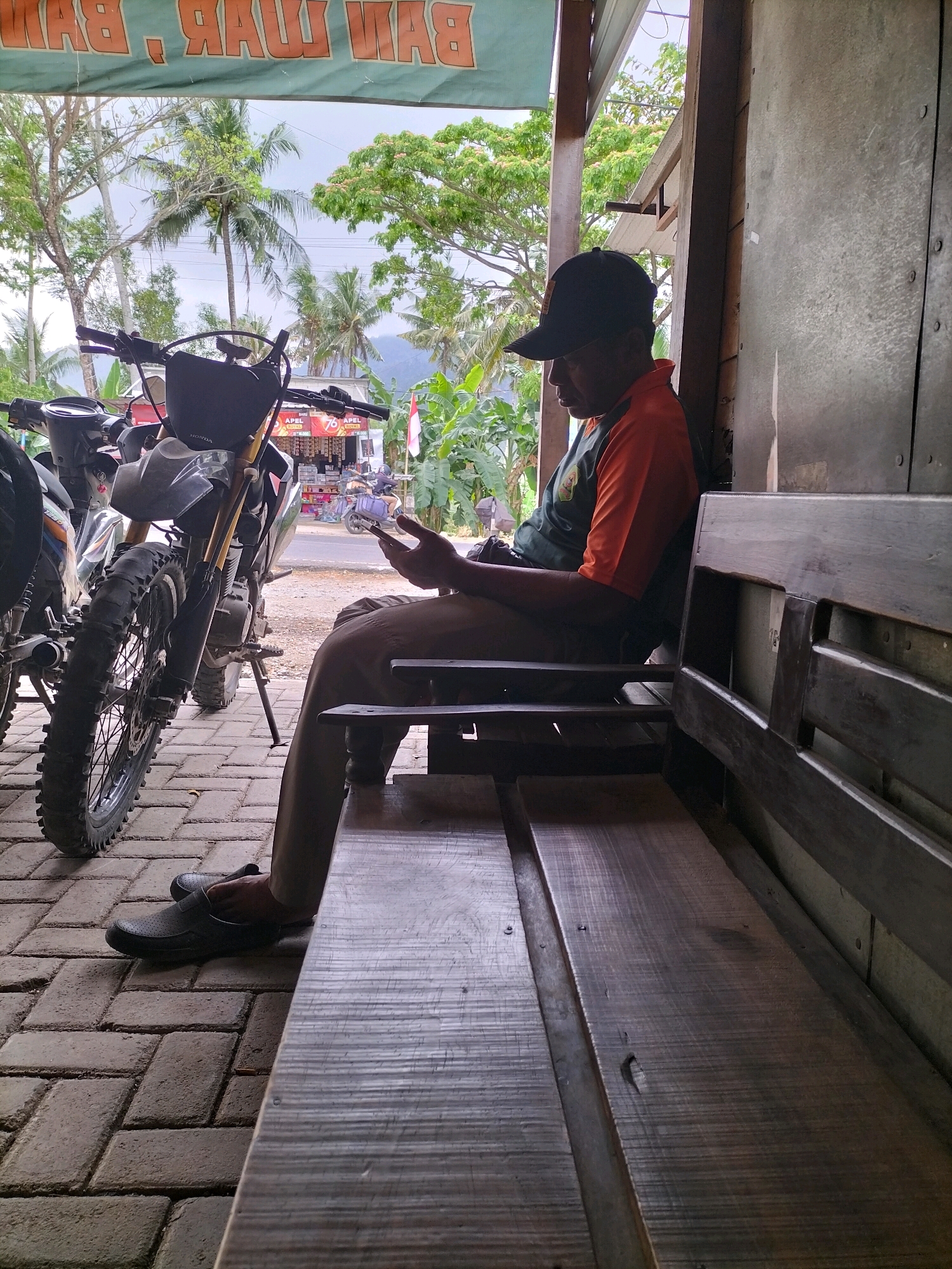 A man wearing a black baseball cap and an orange and green shirt is sitting on a wooden bench, looking at his smartphone. He is wearing brown pants and black slip-on shoes. To his left, a dirt bike is parked on a brick pathway. In the background, a street with buildings and trees can be seen, with mountains in the distance. The scene appears to be in Tasikmadu, Indonesia.