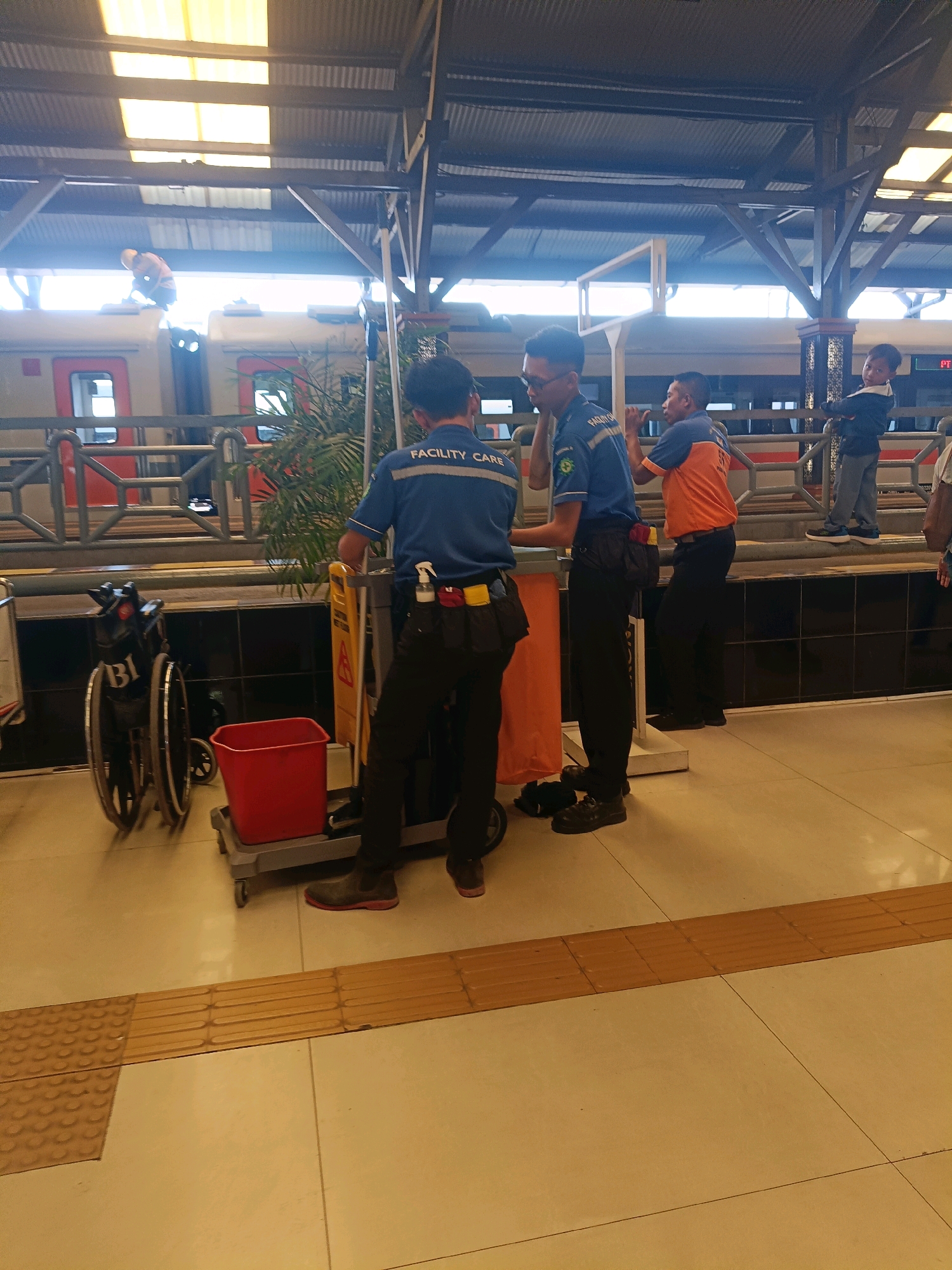 This image depicts a scene at a train station, likely in Surabaya, Indonesia, based on the context. In the foreground, two men in uniform are standing near a cleaning cart. One man, on the left, is wearing a blue shirt with "FACILITY CARE" printed on it and black pants. He is facing away from the camera, engaged with the cleaning cart which holds a red bucket and cleaning supplies. The second man, to his right, is wearing an orange and blue shirt and black pants, and appears to be talking to the first man. Behind them, a train with orange and white livery is visible at the platform. Further in the background, a young boy in a blue jacket and grey pants stands with his arms crossed, observing the scene. Another individual, wearing a blue jacket and grey pants, is visible in the far background near the train. A wheelchair is parked to the left of the cleaning cart. The environment is an open-air train station with a corrugated metal roof supported by beams. The floor is tiled, with tactile paving strips indicating a pathway for the visually impaired. Sunlight filters through the roof, creating bright patches on the platform.