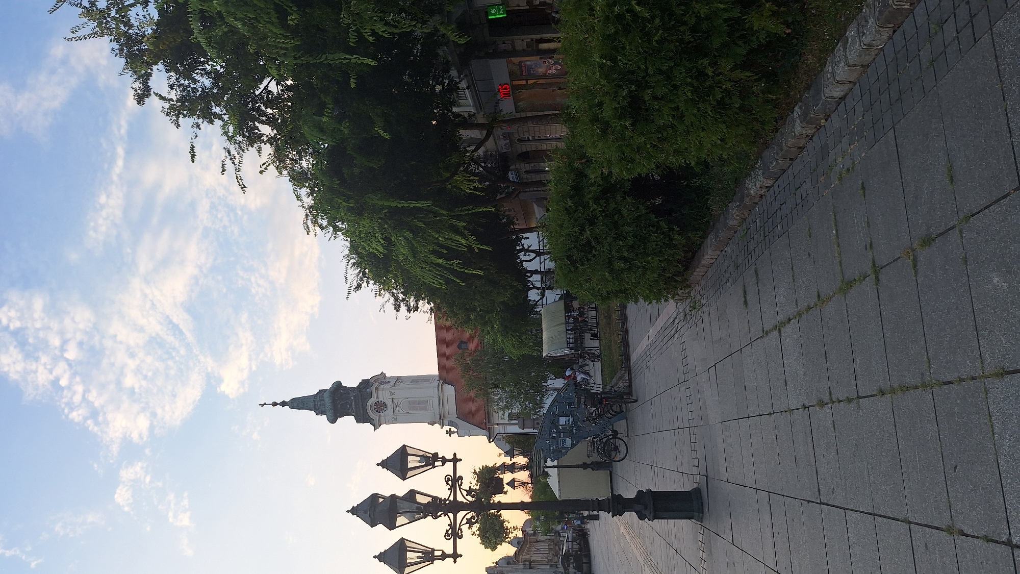This is a ground-level, eye-level, full shot taken from a city street in Sombor, Serbia, capturing an outdoor scene during the day. In the foreground, a large, ornate black lamppost with four lights stands on a paved sidewalk. The sidewalk is made of large, grey square tiles with grass growing in the gaps. To the right of the sidewalk are well-maintained green bushes.
Behind the bushes, a street runs horizontally, lined with trees and buildings. Several bicycles are parked near a white tent structure. Further back, a white church tower with a green steeple and a cross rises above the trees and red-tiled roofs of other buildings. The sky above is a light blue with scattered white clouds. The overall impression is of a pleasant, urban public space.