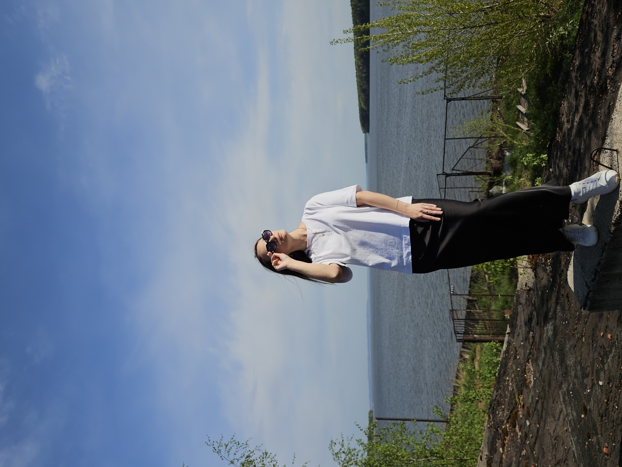 A woman in sunglasses, a white shirt, and a black skirt stands on a concrete structure beside a body of water. She is facing the camera and holding her sunglasses. The water extends to the horizon, with a wooded shoreline in the distance. The sky is blue with some clouds. The concrete structure is worn and partially covered with vegetation. The location is most likely the city of Chaikovsky, Russia.