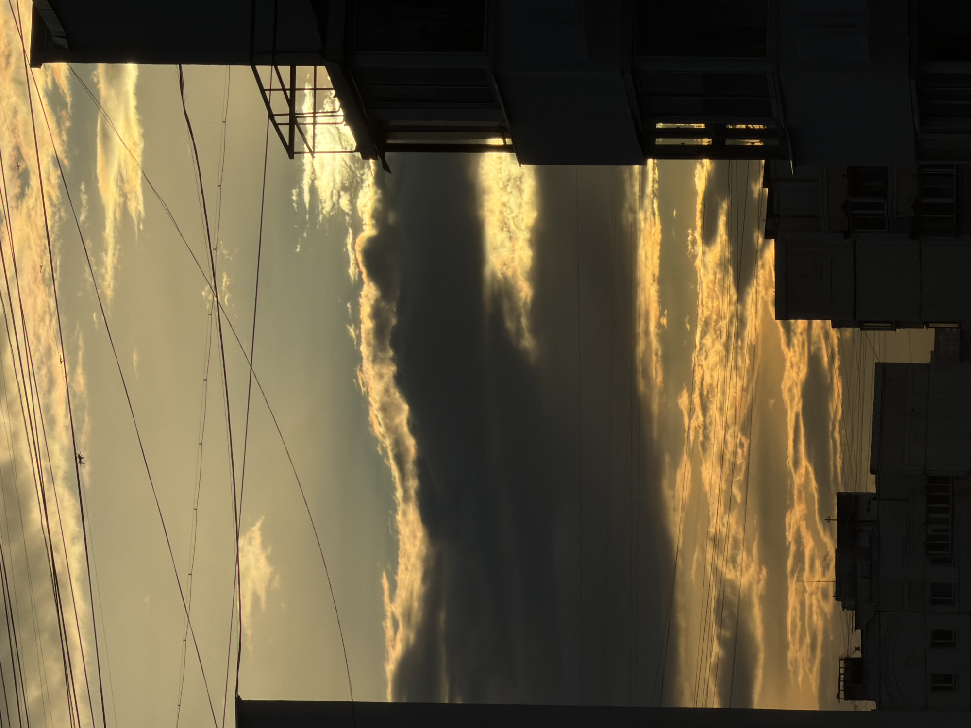 This is a medium shot, taken from a low angle, of the sky and buildings in Khimki, Russia, during sunset. The sky is filled with dramatic clouds that are illuminated by the setting sun, creating a striking contrast between the bright orange and yellow hues of the clouds and the darker, silhouetted buildings. Several power lines are visible, stretching across the frame and adding to the urban landscape. In the foreground, the dark silhouettes of apartment buildings are visible. The building on the right side of the frame has a balcony with railings. The lighting suggests that it is either early morning or late evening. There are no people or animals visible in the image.