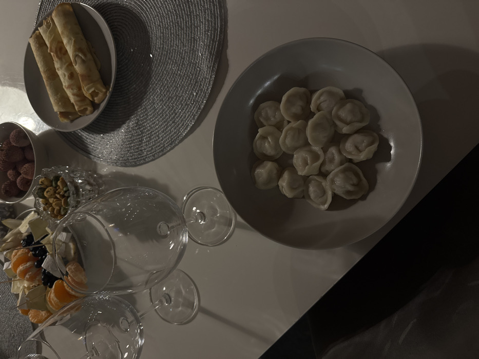 A high-angle shot captures a dimly lit table arrangement. A gray woven placemat supports a plate of rolled crepes. Nearby, a bowl of lychees and a crystal dish of nuts accompany a platter of cheese and tangerine slices. Two wine glasses sit on the table's surface. A separate bowl contains dumplings, positioned beside the glasses. Part of a dark surface is present in the right lower corner.