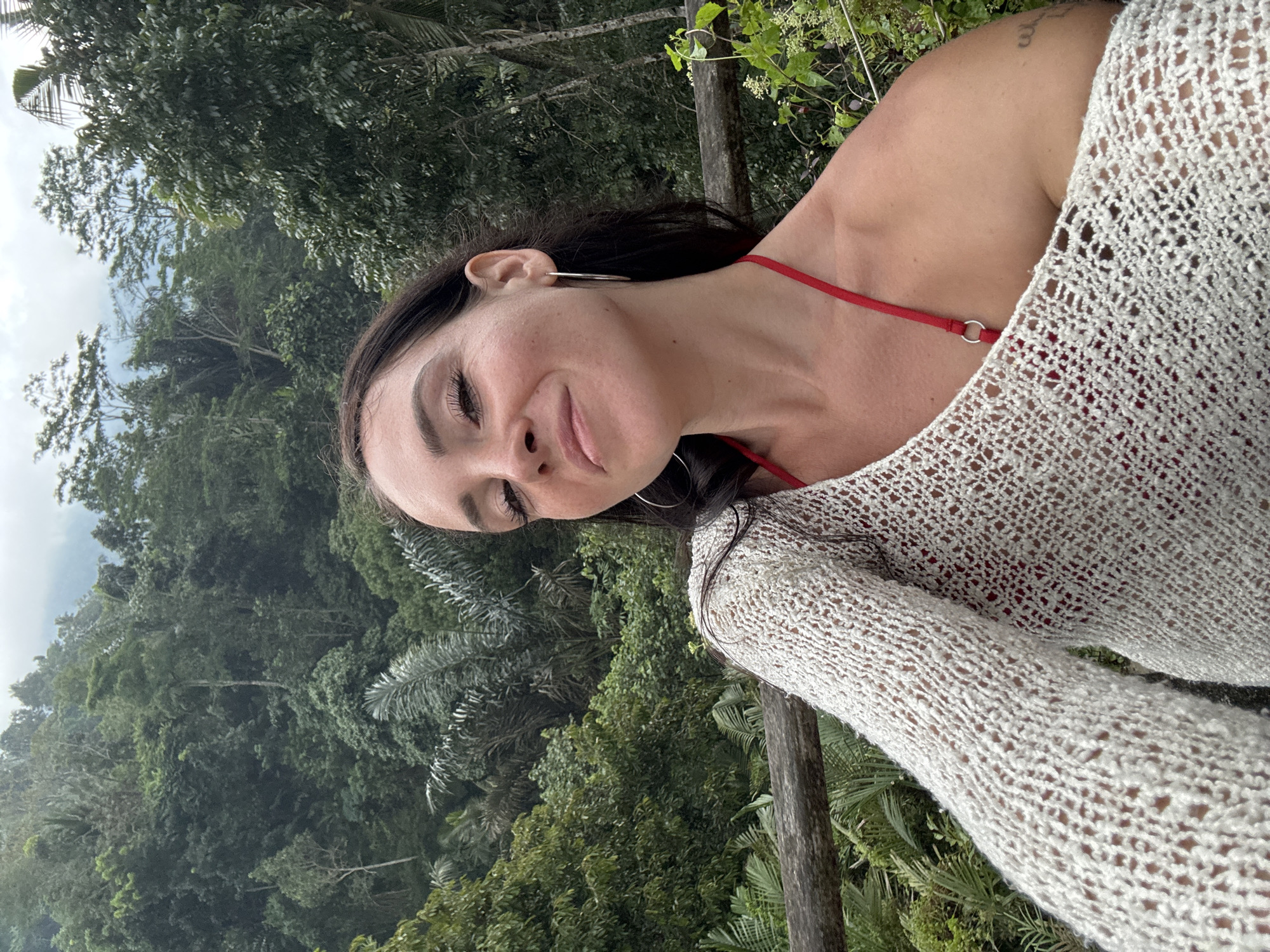 In Bebandem, Indonesia, a woman with dark hair and fair skin takes a selfie outdoors. She is wearing a white knitted cover-up over a red bikini top. The woman is smiling and looking towards the camera. Behind her, a lush green jungle and misty mountains can be seen. The image is a close-up, with the woman's face and upper body filling most of the frame. The natural environment suggests a tropical location, and the mist in the background hints at a cloudy or humid day.