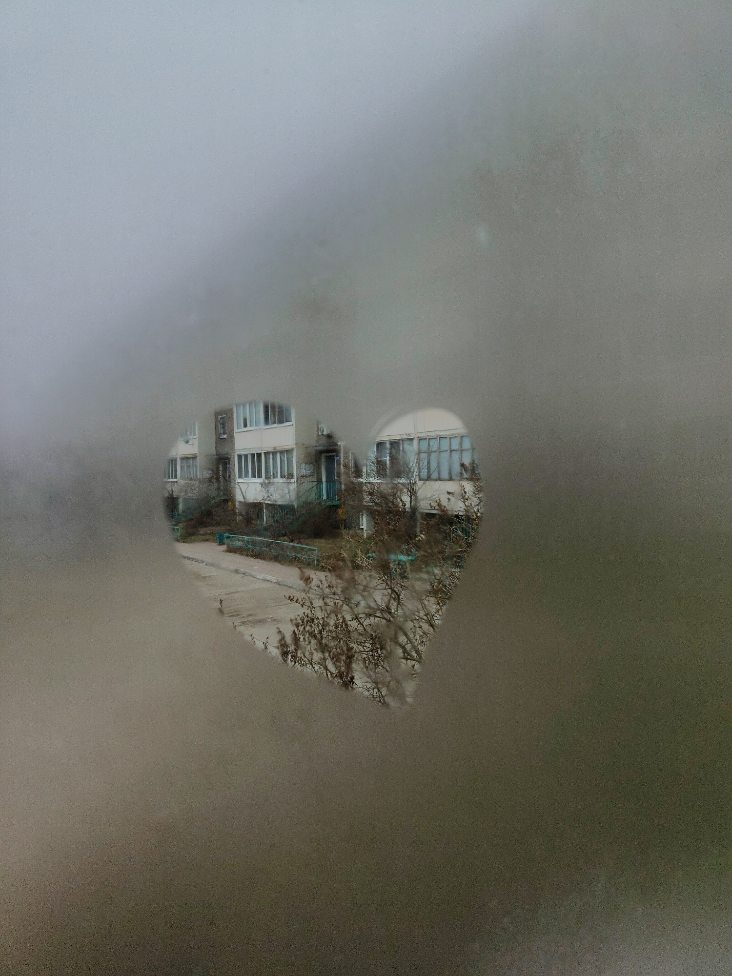 The image shows a view through a partially fogged or frosted window. A heart-shaped area in the fogging has been cleared, providing a view of a multi-story apartment building. The building has a light-colored exterior and multiple windows. There is a sidewalk or paved area in front of the building, and some bare trees or bushes are visible in the foreground. The location appears to be a residential area, potentially an urban or suburban setting, with the city and country Unknown.