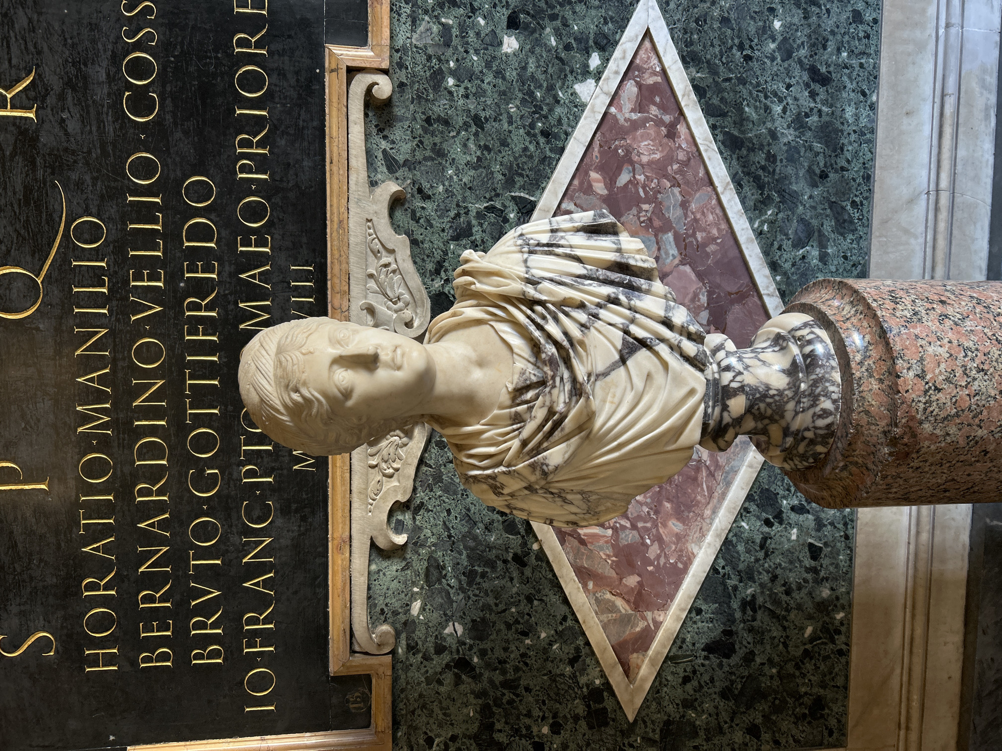 A white marble bust of a woman, sculpted in a classical style, is centrally positioned on a variegated marble base and a cylindrical, mottled reddish-brown granite pedestal. The bust features a draped garment across the shoulders, exhibiting prominent grey veining within the white marble. Behind the bust, a wall is paneled with multiple types of polished marble. The upper section of the wall features a black marble plaque with gilded Latin inscriptions, which include "S P Q R," "HORATIO MANILIO," "BERNARDINO VELLIO COSS," "BRVTO GOTTIFREDO," and "IO FRANC PTO MAEO PRIORE." Below this plaque, a large panel of dark green marble with black and white inclusions displays a central rhomboid-shaped inlay of reddish-brown marble, framed by a thin white marble border. Ornate carved white marble scrolls and a simple wooden frame are situated between the inscription plaque and the green marble panel. A light-colored marble baseboard runs along the bottom of the wall. This scene is located within the Palazzo dei Conservatori, part of the Capitoline Museums, on Capitoline Hill in Rome, Italy.