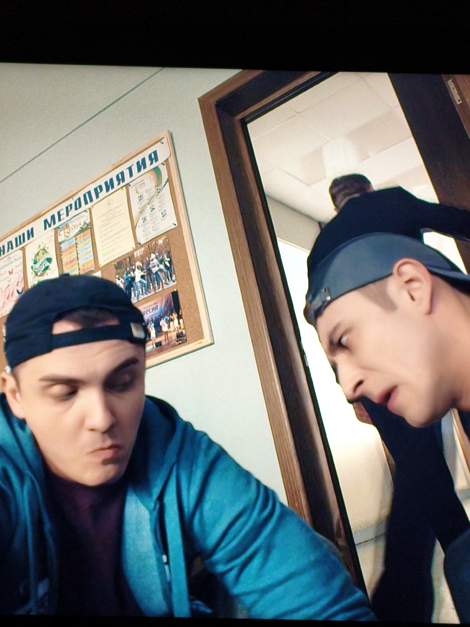 The image depicts two young males indoors, likely in a school or community center setting. In the foreground, a male wearing a teal hoodie and a dark baseball cap is looking downwards with a slightly pensive expression. To the right, partially visible through a doorway, is another male also wearing a dark baseball cap and a dark jacket, facing away from the camera and looking into a brightly lit room. Behind the first male, a bulletin board is visible, adorned with posters and images, some appearing to be event announcements or photographs of performances. The bulletin board has text in Russian, including "НАШИ МЕРОПРИЯТИЯ" (Our Events). The overall environment suggests an informal gathering or an educational institution. There is no indication of the city of Vladivostok, Russia, in the image itself.