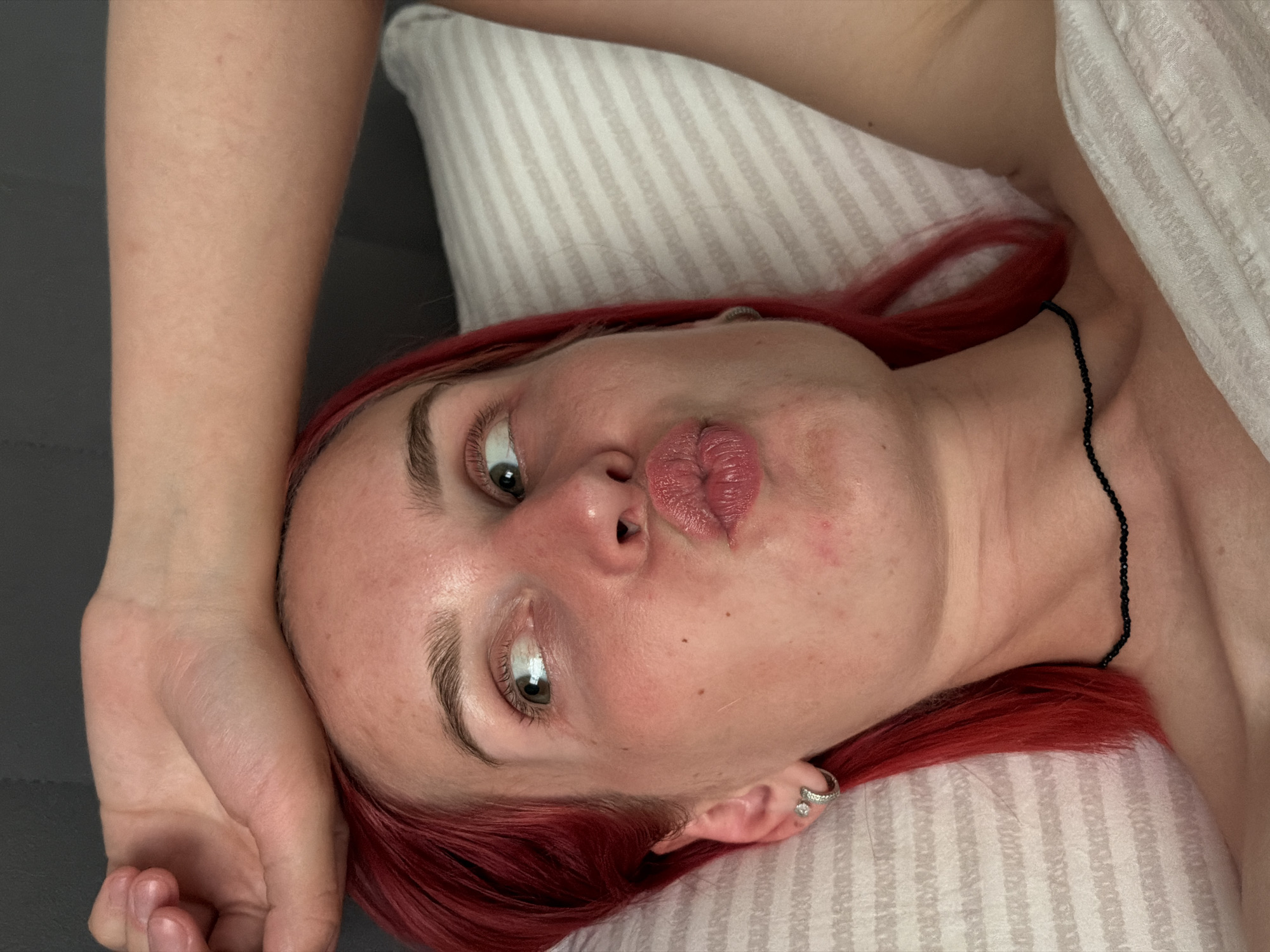 A young woman with bright red hair lies on her side on a striped pillow, looking off to the side. Her right arm is raised and bent behind her head, with her hand resting on her forehead. She is wearing a black beaded necklace. The background appears to be a dark gray surface. The scene is indoors, in what appears to be a bedroom. The location is not specified in the image or the provided information.
