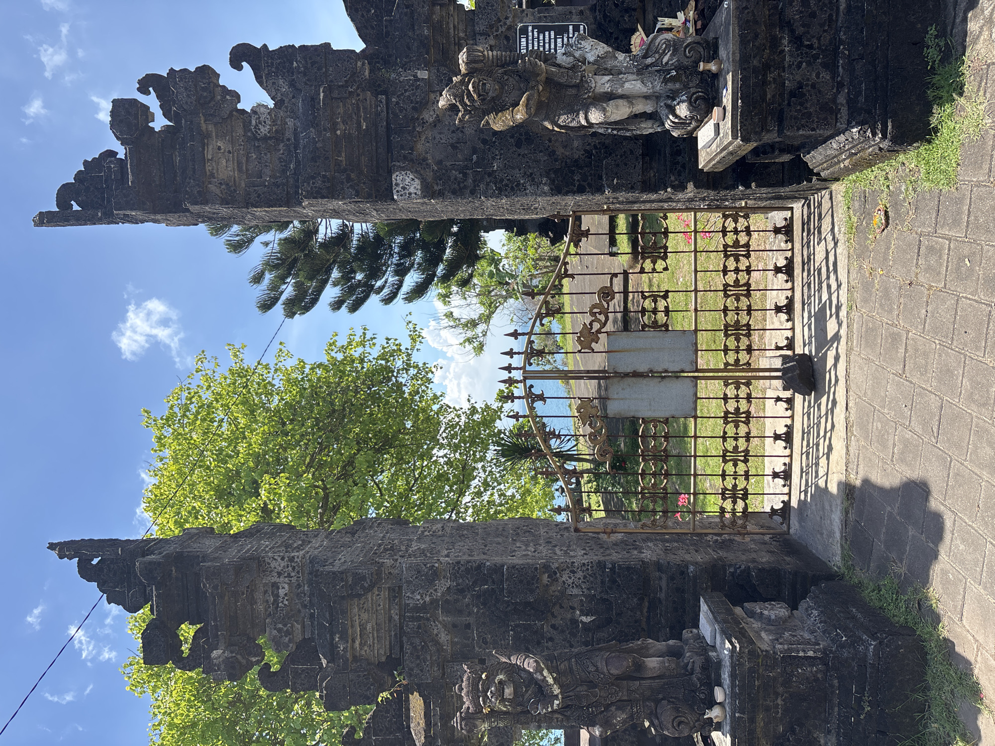 The image shows a view through an ornate, weathered stone gate with intricate carvings, likely a traditional Balinese structure, towards a lush, green outdoor area. The gate is flanked by statues on either side, and a rusted metal gate stands in the center, providing a glimpse of trees, grass, and a body of water in the distance under a blue sky with sparse clouds. The ground is paved with stone bricks, and some vegetation grows along the edges of the gate. There is a sign mounted to the gate. The location is likely in Unknown, Indonesia, based on the Balinese architectural style.