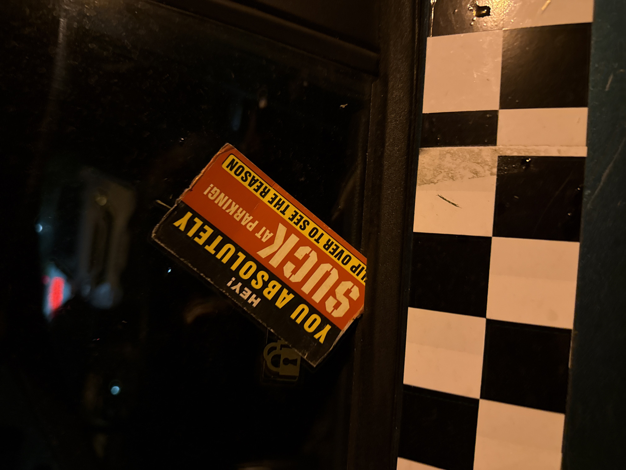 The image depicts a sticker adhered to a window or door in Unknown, Unknown. The sticker is rectangular and displays the message "HEY! YOU ABSOLUTELY SUCK AT PARKING! FLIP OVER TO SEE THE REASON". The sticker is orange with black text and a white border. A small padlock design is visible below the text. To the right of the window or door is a black and white checkered pattern. The overall lighting suggests the scene is either indoors or it is nighttime.