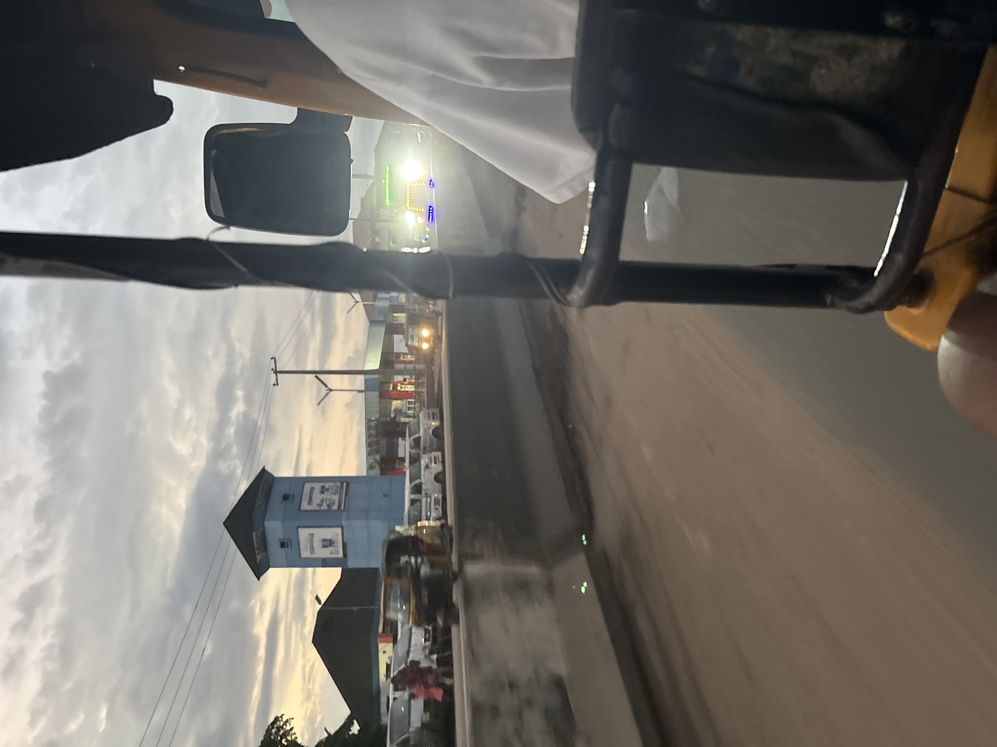 The image depicts a street scene in Onitsha, Nigeria, captured from the perspective of a moving vehicle, likely a rickshaw or a similar three-wheeled transport. The sky is overcast with scattered clouds, hinting at either early morning or late afternoon. On the left side of the road, a prominent blue tower with a dark roof stands out, featuring advertisements on its panels. Next to it, a dark-pitched roof structure is visible. Several vehicles, including white minivans and what appears to be a bus, are parked or moving along the road. Pedestrians, including a woman and children dressed in colorful clothing, are visible near the parked vehicles. A light-colored concrete barrier separates the road from the area where the vehicles are stopped. In the foreground, the interior of the moving vehicle is partially visible, with parts of the frame and a side mirror in view. The road surface appears to be made of compacted earth or gravel. Further down the road, another rickshaw is seen, and on the right, a vehicle with bright, multi-colored lights is approaching. The overall impression is of a busy urban environment.