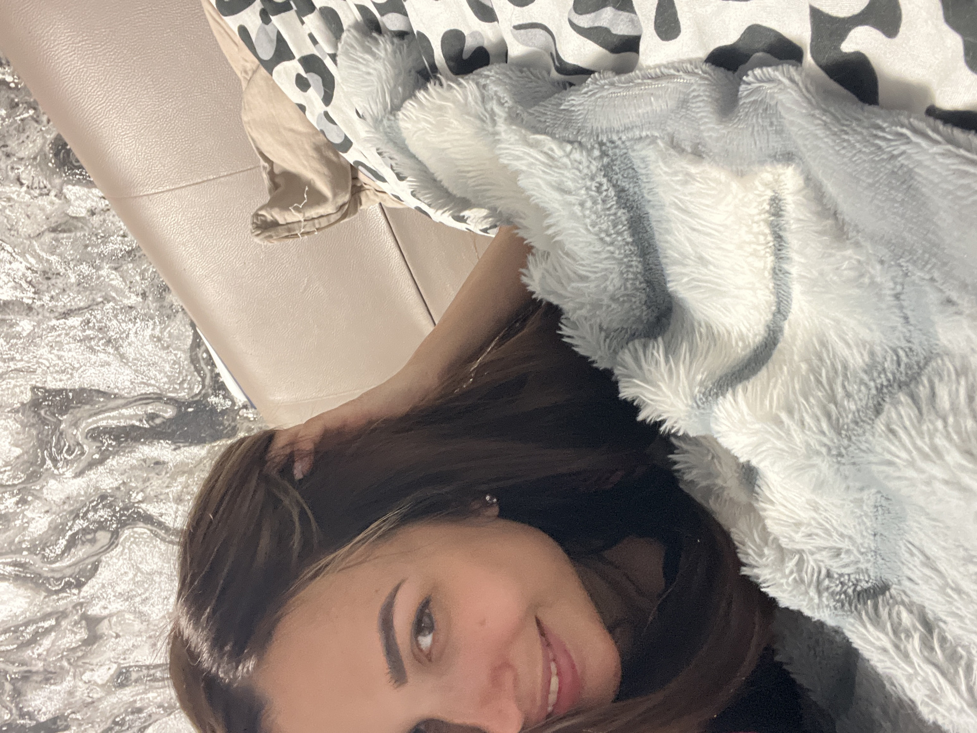 A young woman is photographed from a high angle, lying down. She has long, dark brown hair and is wearing a light-colored, fuzzy blanket that is draped over her. The blanket has a gray, striped pattern. Her right hand is resting on her head, and she is looking at the camera with a smile. Her eyes are brown, and she has dark eyebrows. The background consists of a light-colored couch and a patterned rug. The lighting suggests it could be daytime, but the location is unclear. There is no visible text in the image.