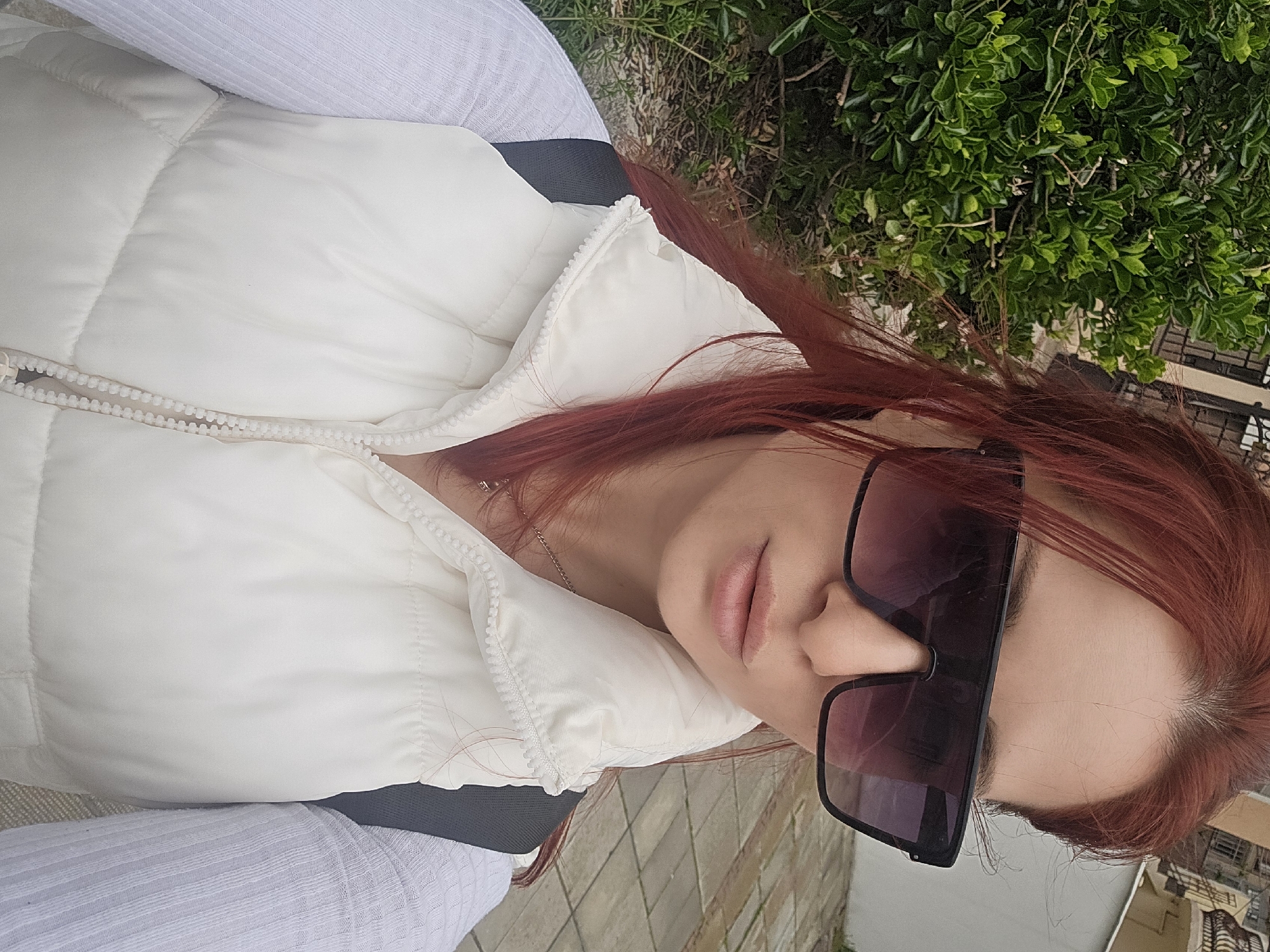 A woman with red hair is wearing a white puffer vest and sunglasses. She is outdoors, likely in Burgas, Bulgaria, indicated by the buildings and greenery in the background. The background suggests a paved area possibly near a residential or commercial building. The image is taken from an upward angle, presenting a close-up of the woman's face and attire.