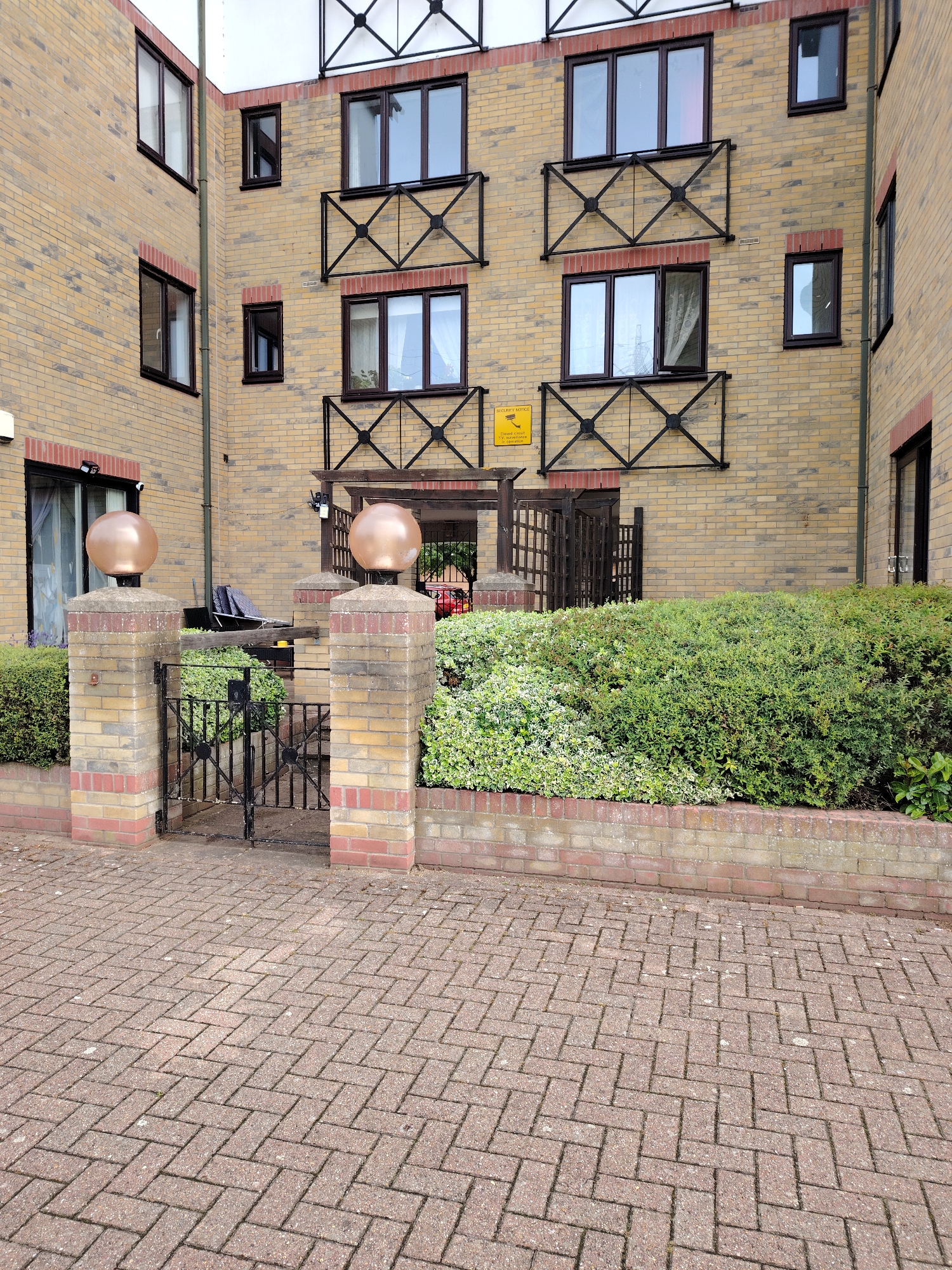 This image depicts the exterior of a multi-story residential building, presumably located in Peterborough, United Kingdom, under overcast daylight conditions. The building's facade is constructed primarily from light brown or yellow bricks, with courses of red bricks accentuating window sills and lintels, as well as outlining a recessed section on the upper floor. Dark-framed windows are symmetrically arranged, with two upper windows featuring dark metal railings below them, characterized by a central 'X' design. Below these, another set of two dark-framed windows also has similar 'X'-patterned metal railings; the rightmost window in this pair is noticeably ajar. A yellow "SECURITY NOTICE - Closed Circuit Television in Operation" sign is mounted on the wall below these lower railings.
At ground level, a dark wooden pergola-like structure covers an entrance. A low, black metal gate, featuring a simpler 'X' pattern, is positioned between two brick pillars. These pillars match the building's brickwork and are topped with large, spherical, metallic-bronze finials. Lush, green shrubbery and hedges, including some variegated plants with lighter green and white leaves, are neatly trimmed along the base of the building and within the gated area. A dark outdoor chair or recliner is partially visible behind the left pillar. Two small white security cameras are mounted above a ground-floor window to the left of the gate. The foreground consists of red-brown interlocking brick paving laid in a herringbone pattern. No people are visible in the scene.