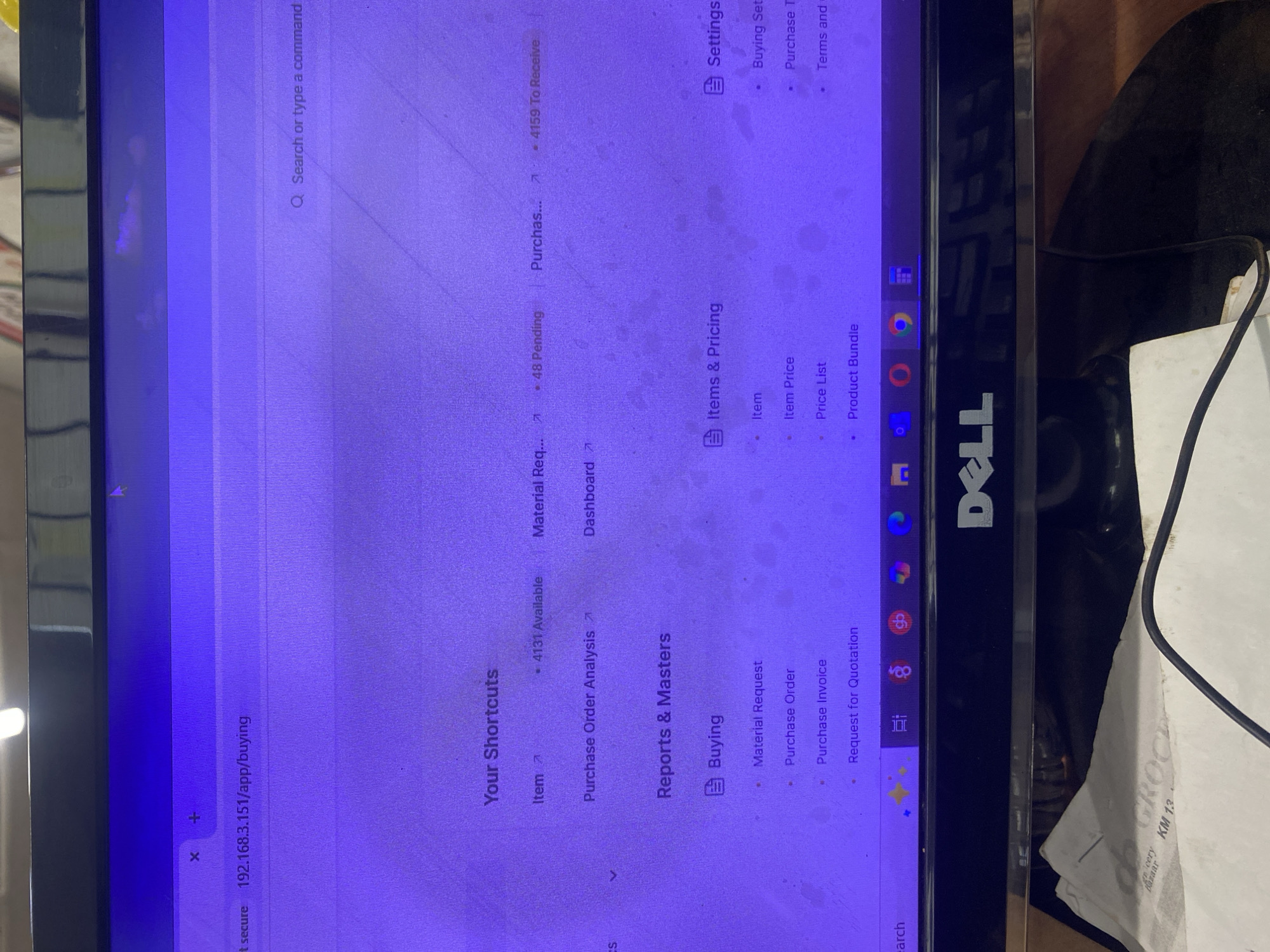 The image shows a Dell computer monitor displaying a web page. The URL visible in the address bar is "192.168.3.151/app/buying". The web page seems to be a dashboard or interface for a business or e-commerce application, with sections such as "Your Shortcuts," "Reports & Masters," "Items & Pricing," and "Settings." The "Your Shortcuts" section has links for "Item," "Material Request," and "Purchase." Under "Reports & Masters" the category of "Buying" is visible with options for "Material Request," "Purchase Order," "Purchase Invoice," and "Request for Quotation." A piece of paper with the text "GROCERY KM 13" is resting on a surface next to the monitor. The text suggests a grocery store and the location "KM 13," potentially referring to a kilometer marker on a road, which is likely along a road in Ikorodu, Nigeria.
