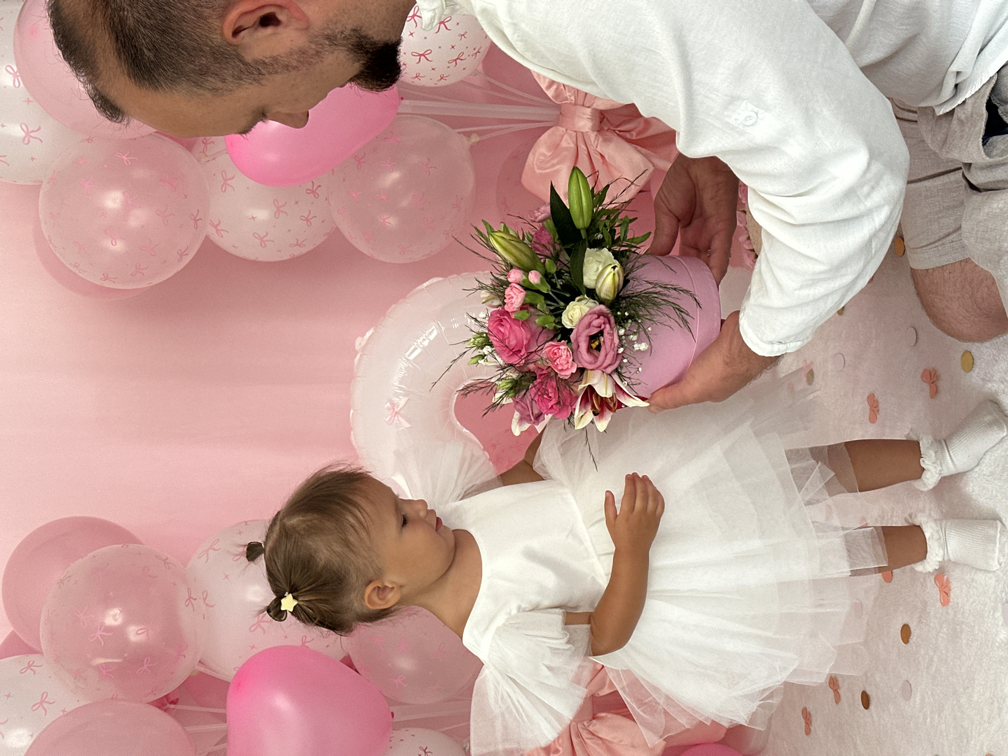 In Wisła, Poland, a man in a white shirt and grey shorts kneels on a white carpeted floor, presenting a pink flower box to a young girl. The girl, wearing a white tutu dress and white socks, stands facing the man, looking down at the flowers. The background is adorned with numerous pink balloons of various sizes, some featuring white bow patterns, and a large pink bow decoration. Confetti pieces are scattered across the floor.