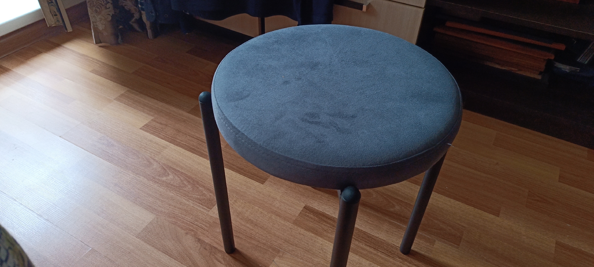 A low-angle shot shows a dark grey, round stool with three thin, dark legs on a wooden floor. The stool is centrally located in the frame, and its top surface is slightly worn. The floor is made of light brown wood planks. In the background, a dark wooden shelf unit is visible, containing several stacked items, possibly books or files. A portion of a dark curtain is also visible in the upper left background. The scene appears to be indoors, in a domestic setting. The image does not provide any information or context to determine its location in Cheboksary, Russia.