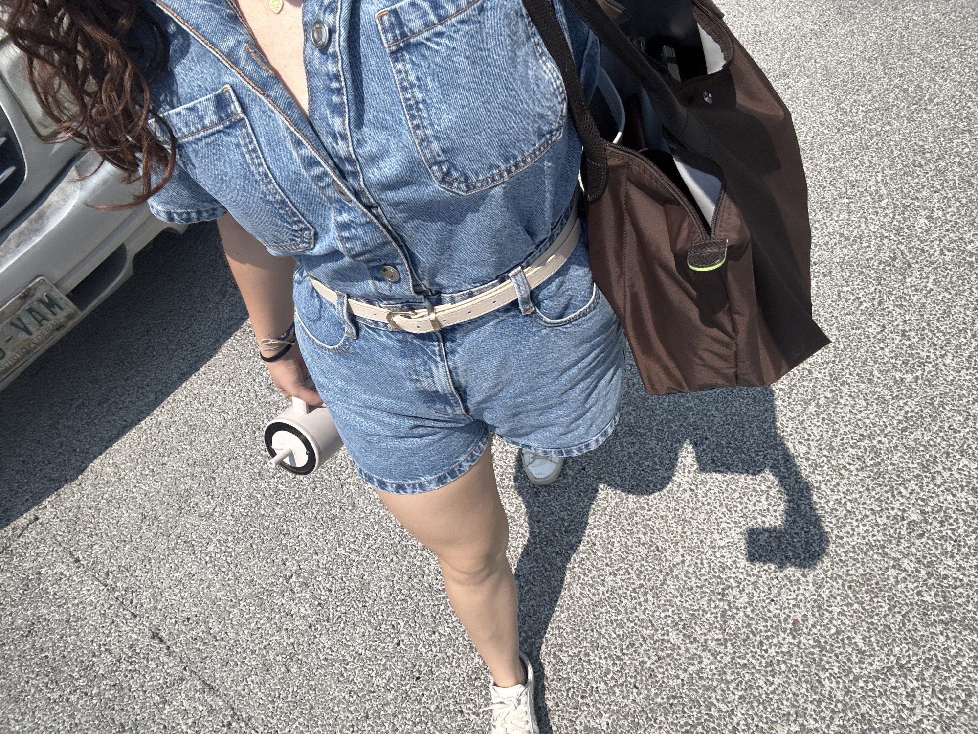 A person in a denim jumpsuit and a light-colored belt is standing on a grey asphalt surface. They are holding a white thermos in their left hand and have a brown tote bag hanging from their right shoulder. The person is wearing white sneakers. A vehicle is partially visible in the background on the left, with license plates labeled "DISTRITO FEDERAL" and "-VAM". The setting appears to be an outdoor paved area in Mexico City, Mexico.