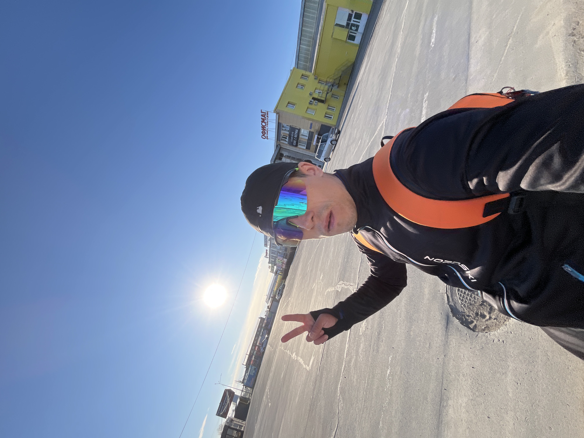 A man wearing sunglasses and an orange backpack is taking a selfie on a paved area. He is giving a peace sign. In the background is a yellow building with a sign that reads "ОФИЦИАТ" (Officiat). The photo was taken in Chaikovsky, Russia.