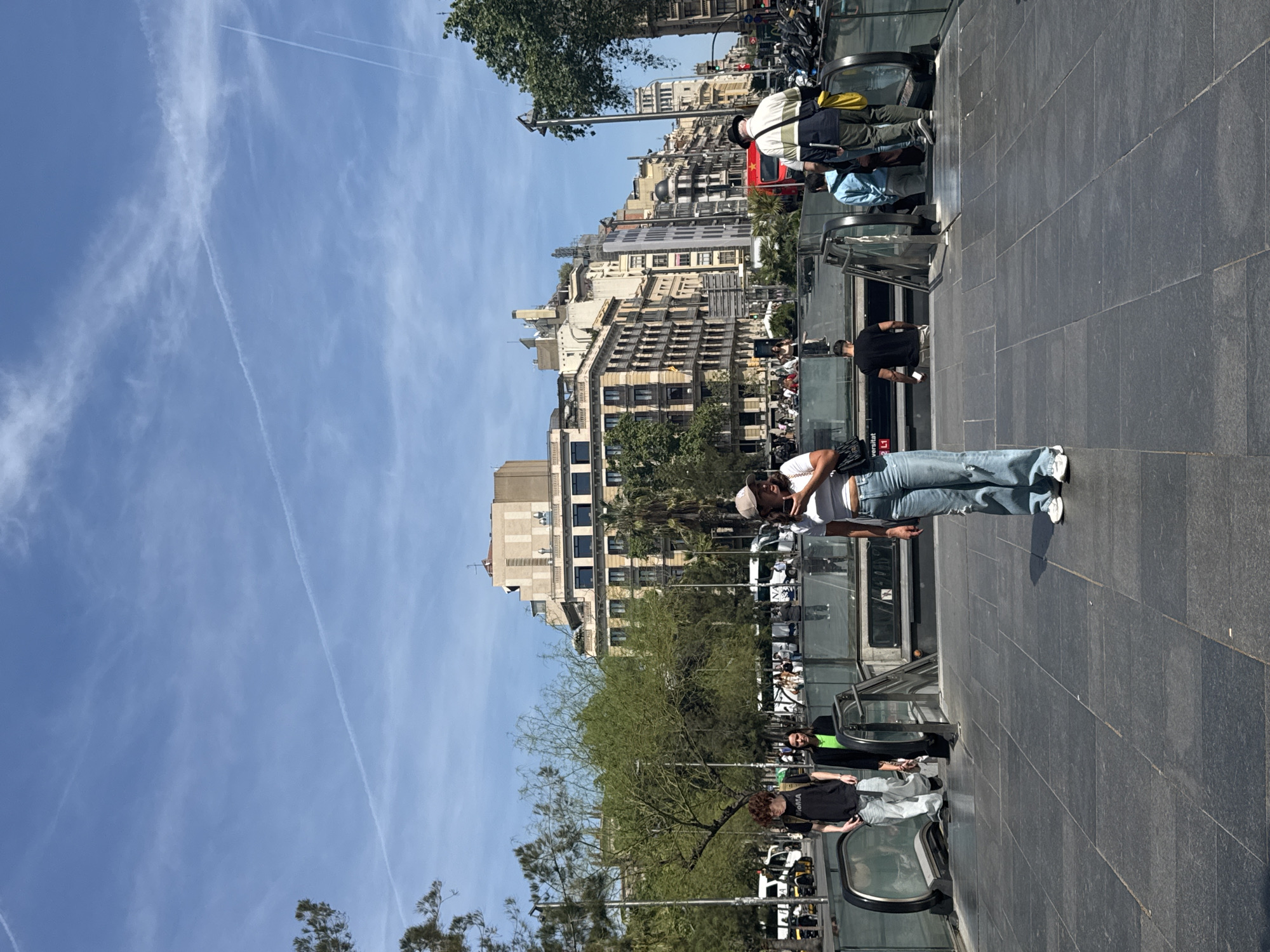 This image depicts a sunny urban street scene in Barcelona, Spain, specifically around a metro station entrance. The time of day is bright daylight, evidenced by clear shadows and the vibrant blue sky which features wispy white clouds and several distinct airplane contrails.
In the mid-ground, a woman wearing a white t-shirt, light blue baggy jeans, and a light-colored cap stands on a dark grey paved surface, facing towards the left side of the frame as if looking down towards the escalators. To her left, several escalators lead down into an underground passage or metro station. Various people are using these escalators; one individual in a black and white striped shirt with a yellow backpack is ascending, and another in a light blue jacket is also visible. Further back and to the left, a man in a black t-shirt and light-colored pants stands near another escalator.
The background is filled with multi-story buildings exhibiting classic European urban architecture, featuring numerous windows and balconies. Green trees are interspersed along the street, adding natural elements to the cityscape. Parked motorbikes or scooters are visible further down the street. The ground is paved with large, dark grey square tiles.
A significant location cue is visible text "Universitat L1" on the glass barrier of one of the escalator entrances, indicating the Universitat metro station on the L1 line in Barcelona. Another piece of text, "WOMAA", can be seen on the back of a black t-shirt worn by a person in the mid-ground. The overall scene captures typical daily activity in a bustling city environment.