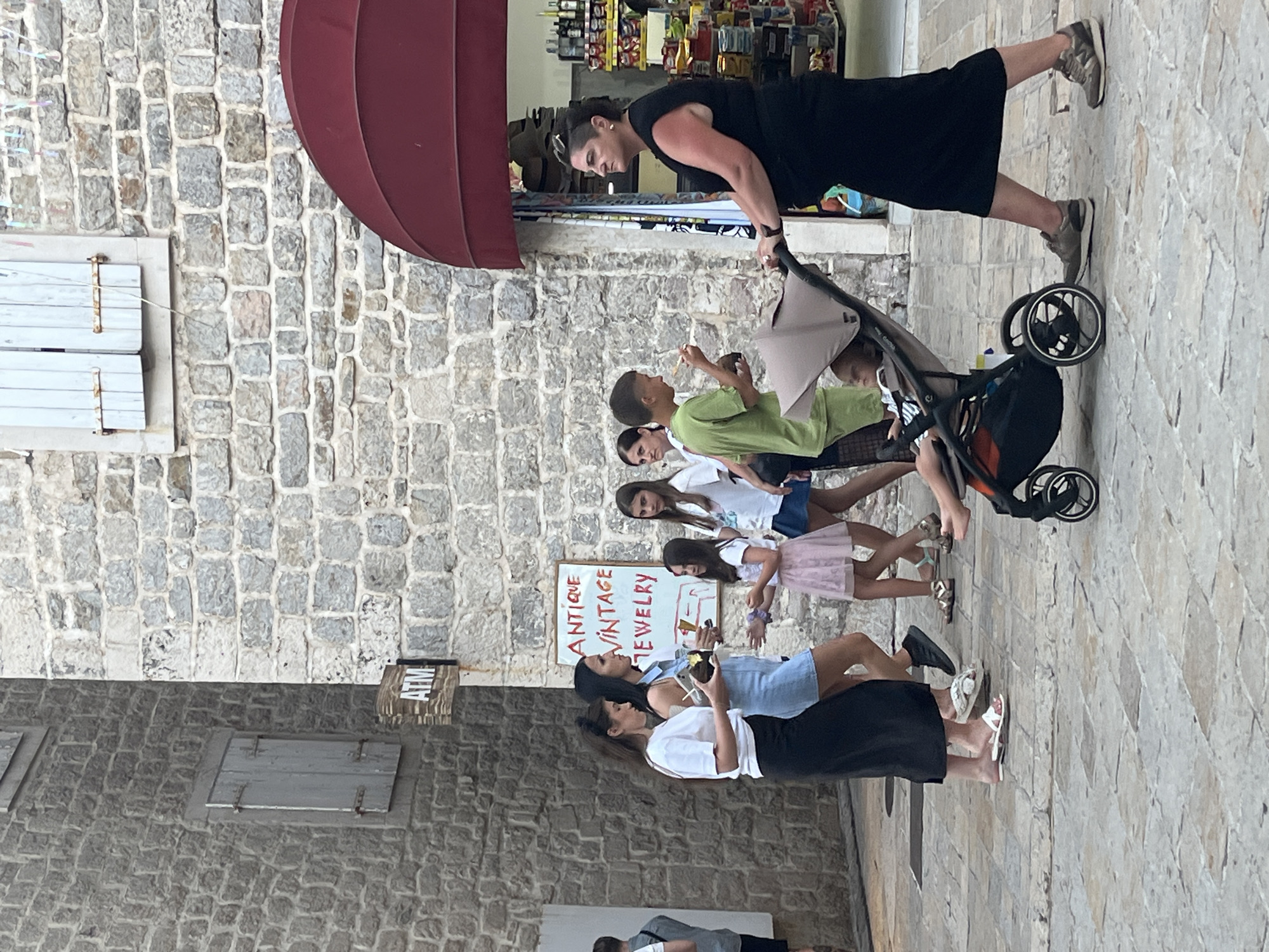 The image depicts a street scene in the old town of Budva, Montenegro, characterized by its stone-paved street and old stone buildings. In the foreground, a woman in a black dress is pushing a stroller containing a baby. To her left, a group of people are walking, including a woman in a white t-shirt and black skirt, another woman in a light blue dress, and several children. Behind this group, a sign reads "ATM." Further to the left, an ATM machine is visible on the stone wall of a building. The background shows more stone buildings, one with wooden shutters. A red awning extends from a shop to the right, displaying various items for sale. The overall atmosphere suggests a warm day, with people strolling through the historic town.