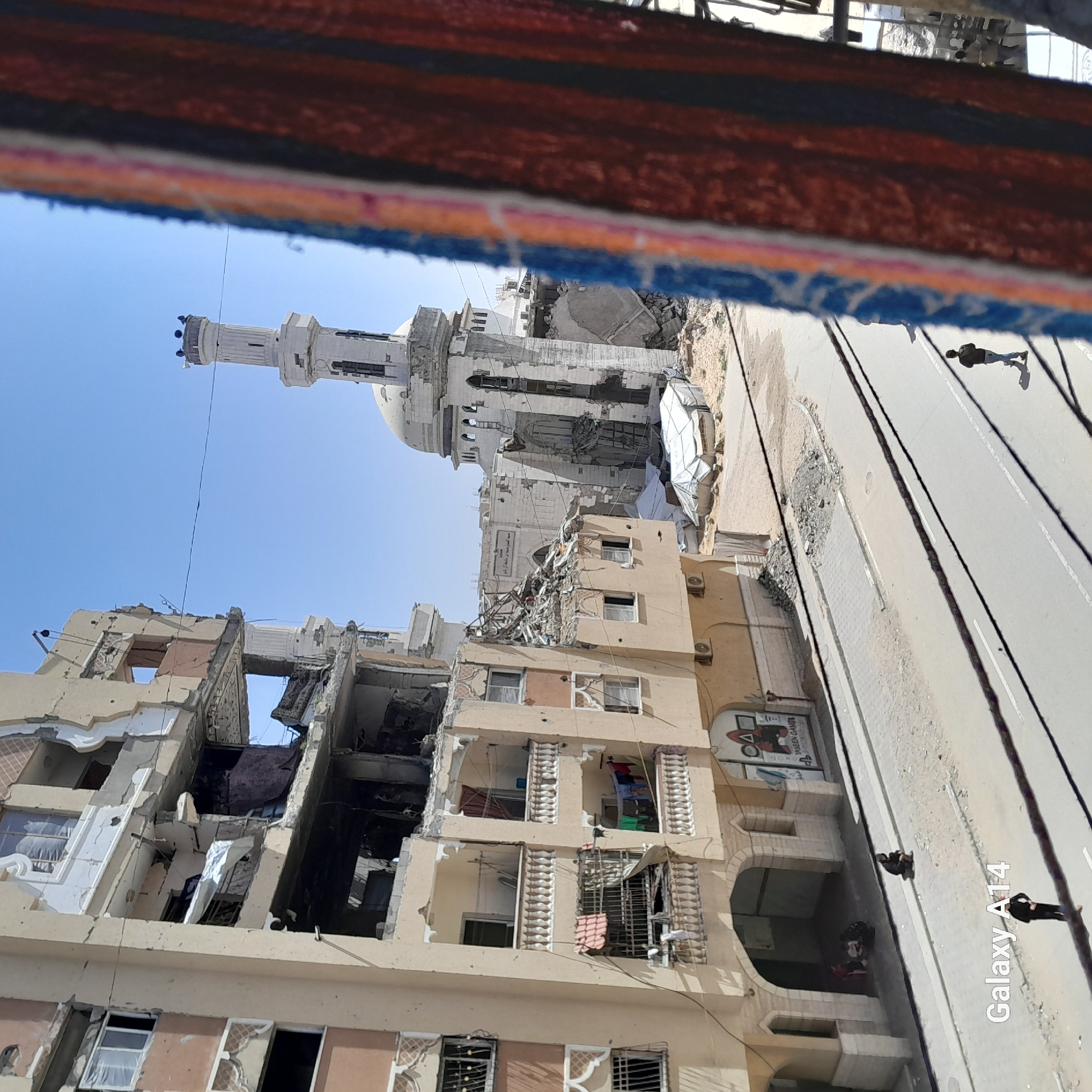 The image depicts extensive damage to buildings and a mosque in what appears to be Khan Yunis, Palestinian Territory. Several multi-story buildings show significant structural damage, with collapsed walls and exposed interiors. A nearby mosque with a tall minaret also exhibits damage. Debris and rubble are visible around the buildings and on the adjacent road. A few people are seen on the street.