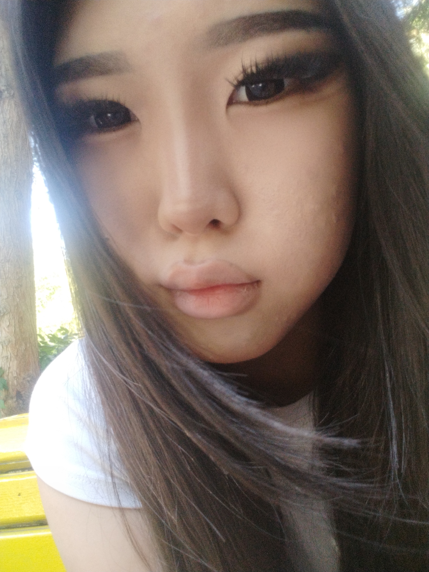 A close-up selfie of a young East Asian woman's face and upper torso is captured outdoors. She has long, dark hair that frames her face and falls over her shoulder. She is wearing a white t-shirt. Her makeup includes dark eyeliner and mascara, with noticeable false eyelashes. Her lips are slightly parted and appear to have a light-colored lip balm or gloss. The background suggests an outdoor setting with trees on the left and hints of greenery. A bright yellow object, possibly a bench, is visible on the lower left side of the frame. The lighting indicates it is daytime, with sunlight creating a bright area to the left of her face. The image has a slightly grainy quality. The specific location in Cholpon-Ata, Kyrgyzstan, is not discernible from the visual information in the image itself; the provided information is an external detail.