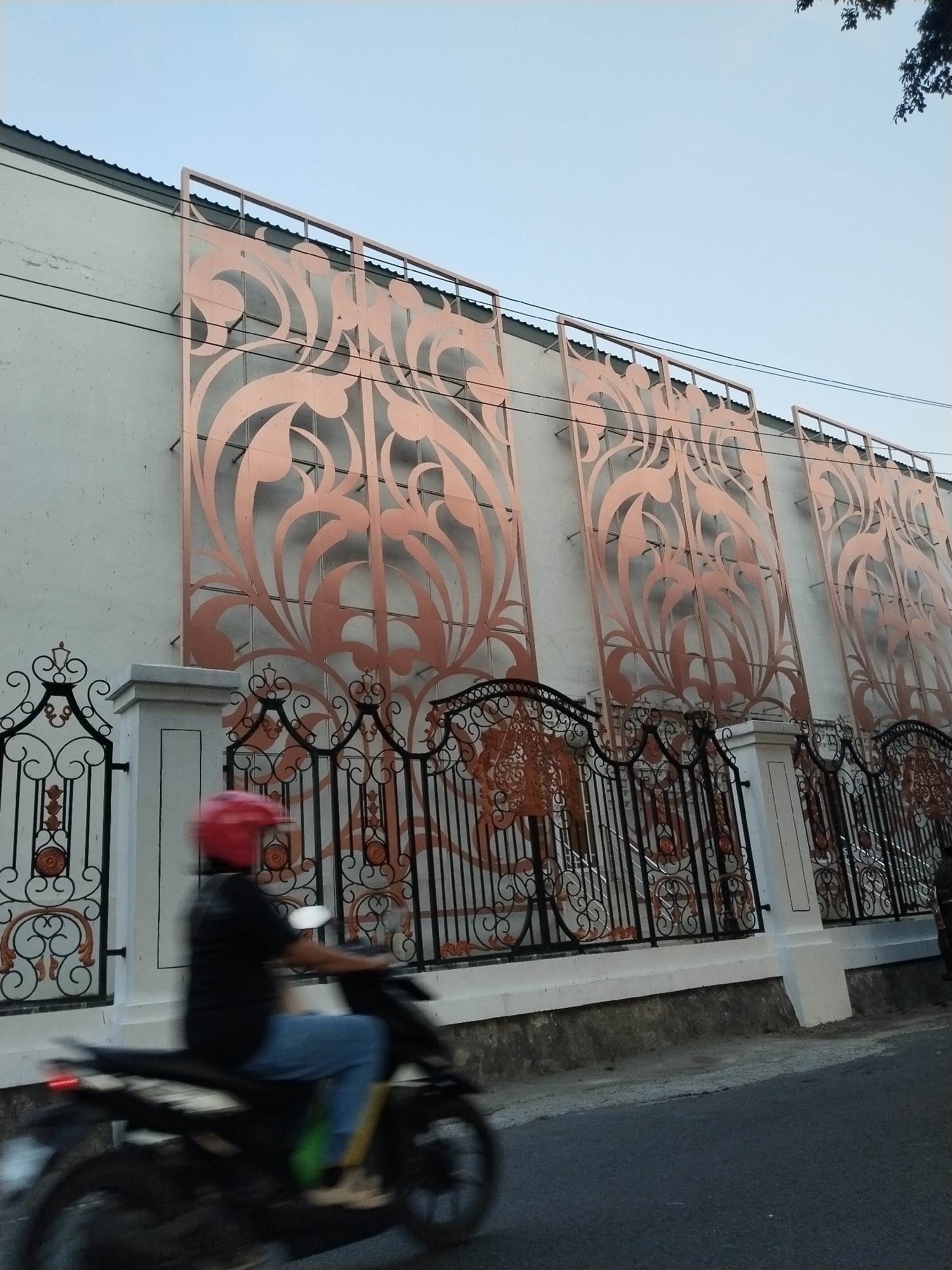 The image depicts a street scene in Sukoharjo, Indonesia. In the foreground, a person wearing a red helmet and blue jeans is riding a motorcycle, their motion blurred. The motorcycle is moving from left to right across the frame. Behind the motorcycle and partially obscuring the background is an ornate black metal fence with copper-colored floral accents. This fence runs along a white wall. Above the wall, large, decorative panels with a swirling copper-colored design are mounted. These panels appear to be made of metal and are attached to a lighter-colored building. The sky is a pale blue, with a sliver of dark tree branches visible in the upper right corner. The overall impression is a decorative building facade and street view under a clear sky.
