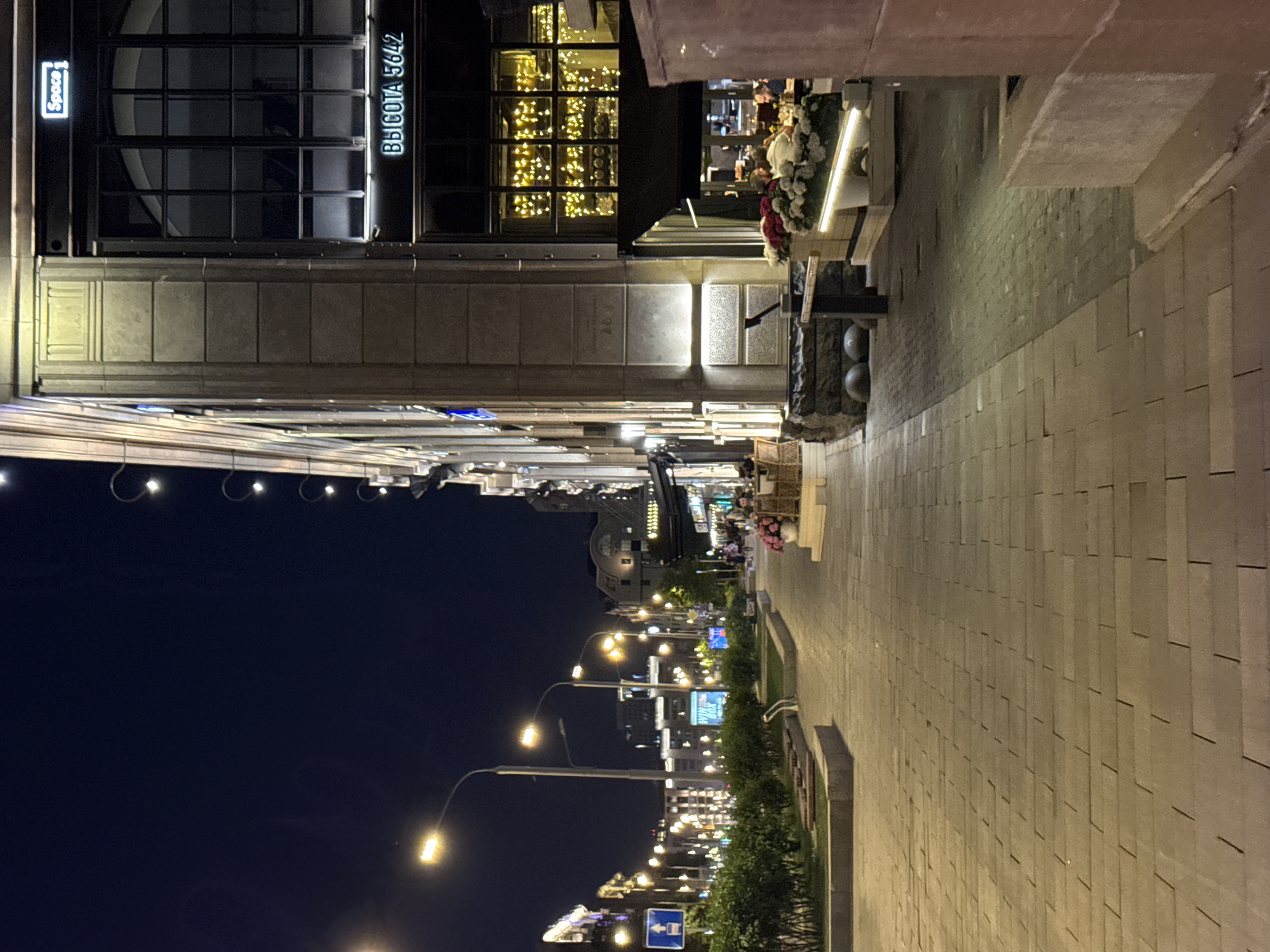 This image depicts a nighttime urban street scene in Moscow, Russia, characterized by artificial illumination. The perspective is from a wide, light-colored paved sidewalk, constructed from rectangular interlocking pavers, extending into the distance.
On the left side of the frame, a row of green bushes separates the sidewalk from a roadway, which is lined with numerous streetlights emitting a warm yellow glow. A blue rectangular sign on a pole displays a white upward arrow over a bus icon, indicating a bus lane or public transport direction. Buildings further down the street on the left are also illuminated. The sky above is dark blue-black.
The right side of the image is dominated by a multi-story building with a facade of light-colored stone or concrete. Near the top of the visible section, a white neon sign reads "Space" above a large, grid-paned window. Below this, another prominent white neon sign displays "ВЫСОТА 5642" (VYSOTA 5642), identifying a restaurant known to be located in Moscow. Beneath this sign, a large window is brightly lit from within by numerous small, warm-toned string lights. An outdoor dining patio is positioned below this window, covered by a dark awning. Several individuals are seated at tables in this area, engaged in dining or conversation. The patio edge is adorned with large planters containing pink and white flowers, which are illuminated from below. Dark, textured stone blocks and smooth, dark spherical elements are visible at the base of the building near the patio, alongside decorative wall-mounted lighting fixtures.