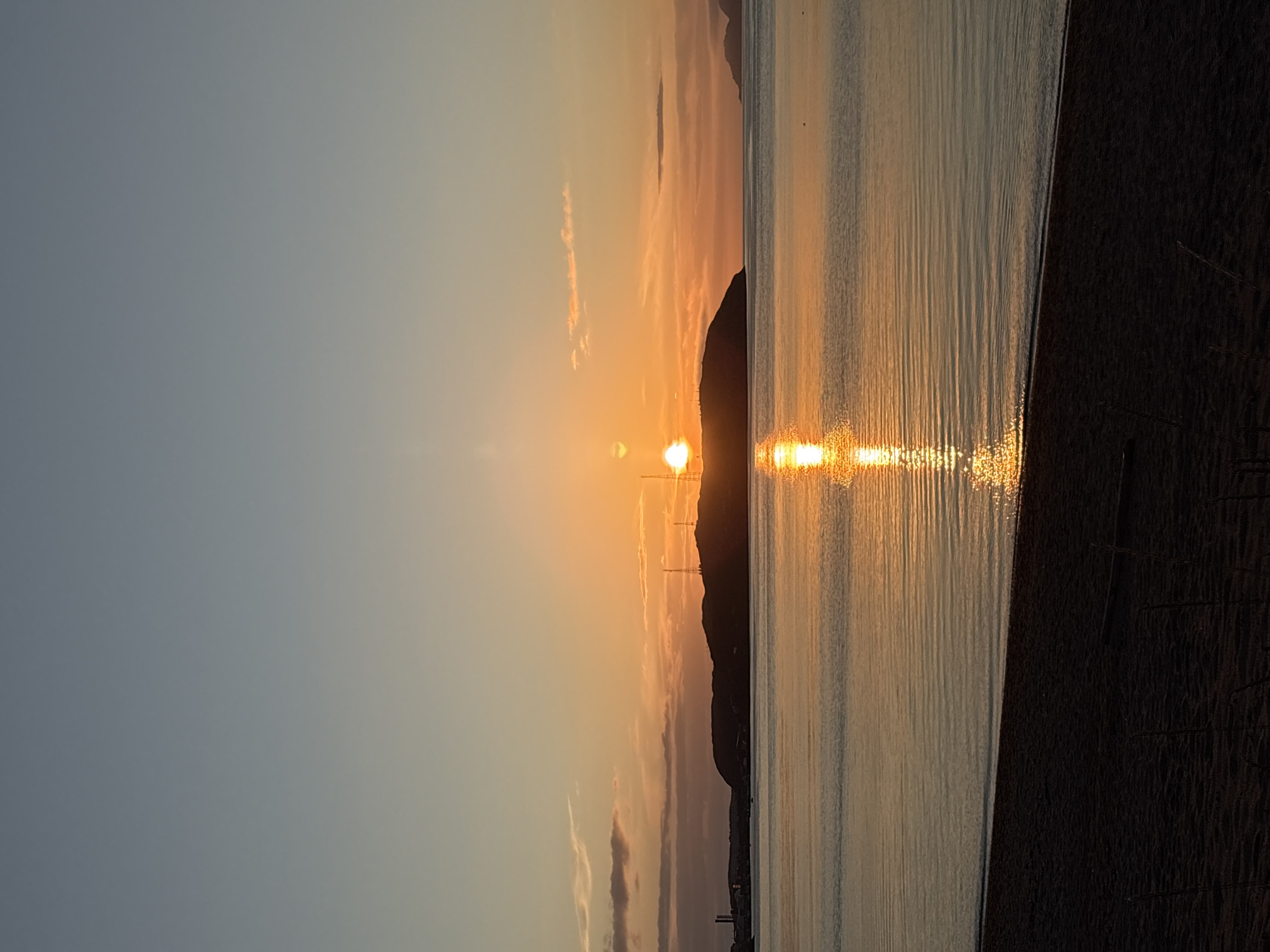 An evening seascape in Teriberka, Russia, features a sunset casting a warm orange glow across the sky and water. The sun, a brilliant orb, is partially obscured by a silhouetted island in the distance. The island is topped with transmission towers. A reflection of the sun shimmers on the calm water, creating a bright path. In the foreground, a dark sandy beach slopes down to meet the water. The sky transitions from a pale blue at the top to a soft orange and pink near the horizon, with a few wispy clouds scattered throughout. The overall scene is peaceful and serene.