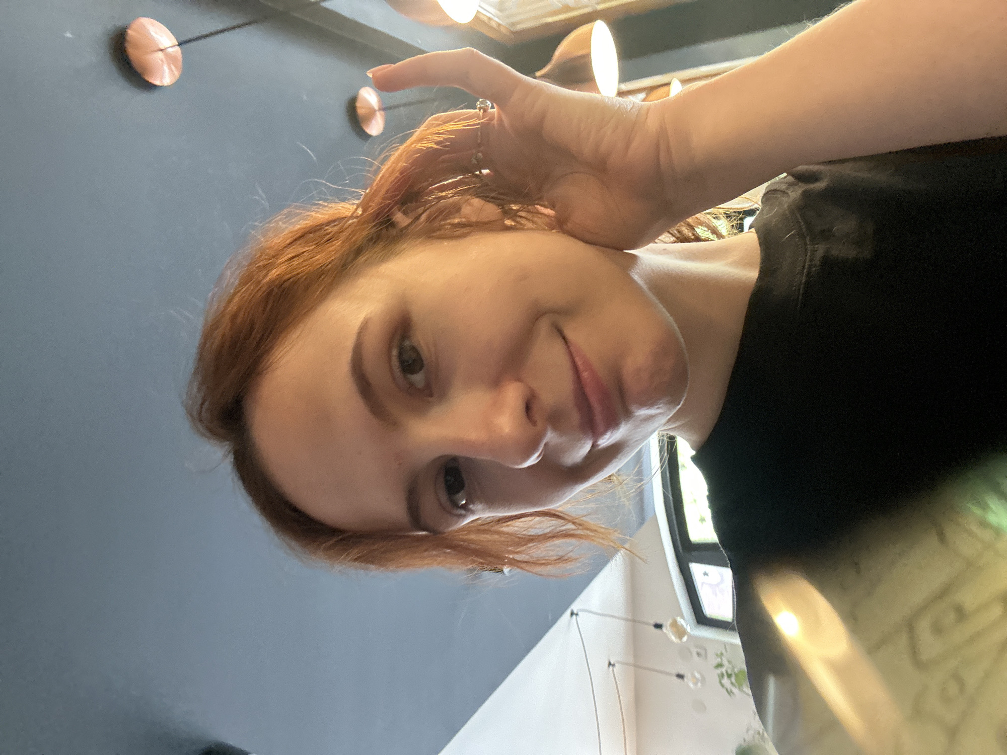 A selfie image shows a light-skinned woman with shoulder-length reddish-brown hair, wearing a black shirt, with her right hand touching her hair. The background consists of a dark blue wall with decorative copper-colored discs, and a window and light fixture are partially visible behind her. Based on the interior design and lighting, it might be taken in a modern cafe or apartment. There is no indication of Novi Sad or Serbia in the image.