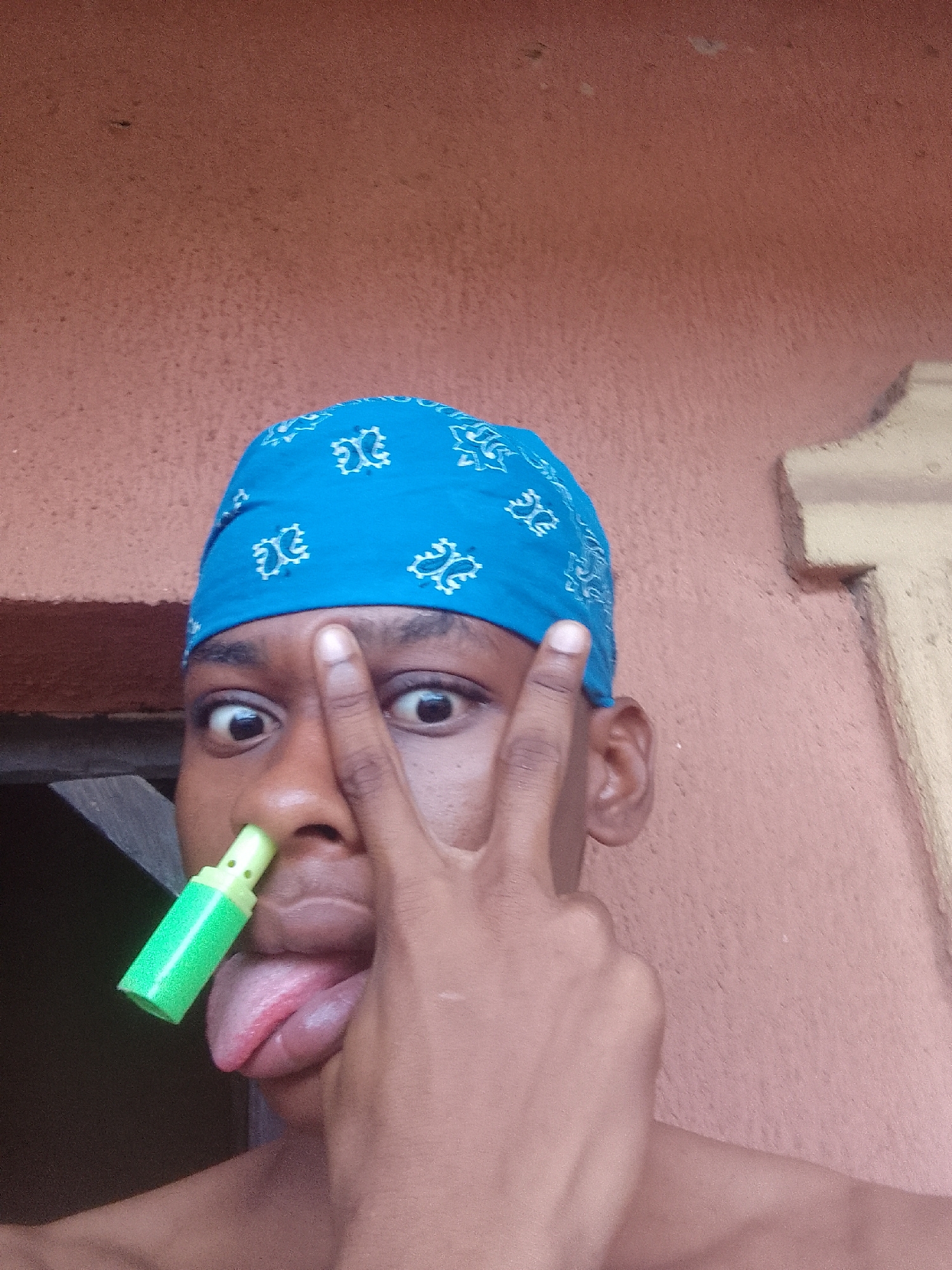 A young African man with dark skin is depicted from the chest up against a textured pink wall. The man wears a blue bandana with a white paisley pattern on his head. He is shirtless. A green plastic object with a lighter green cap is inserted into his left nostril. His tongue is sticking out. His left hand is raised in a peace sign, positioned over his eyes. The setting is a residential area in Nkpor, Nigeria, marked by the pink wall and typical construction details of the region.