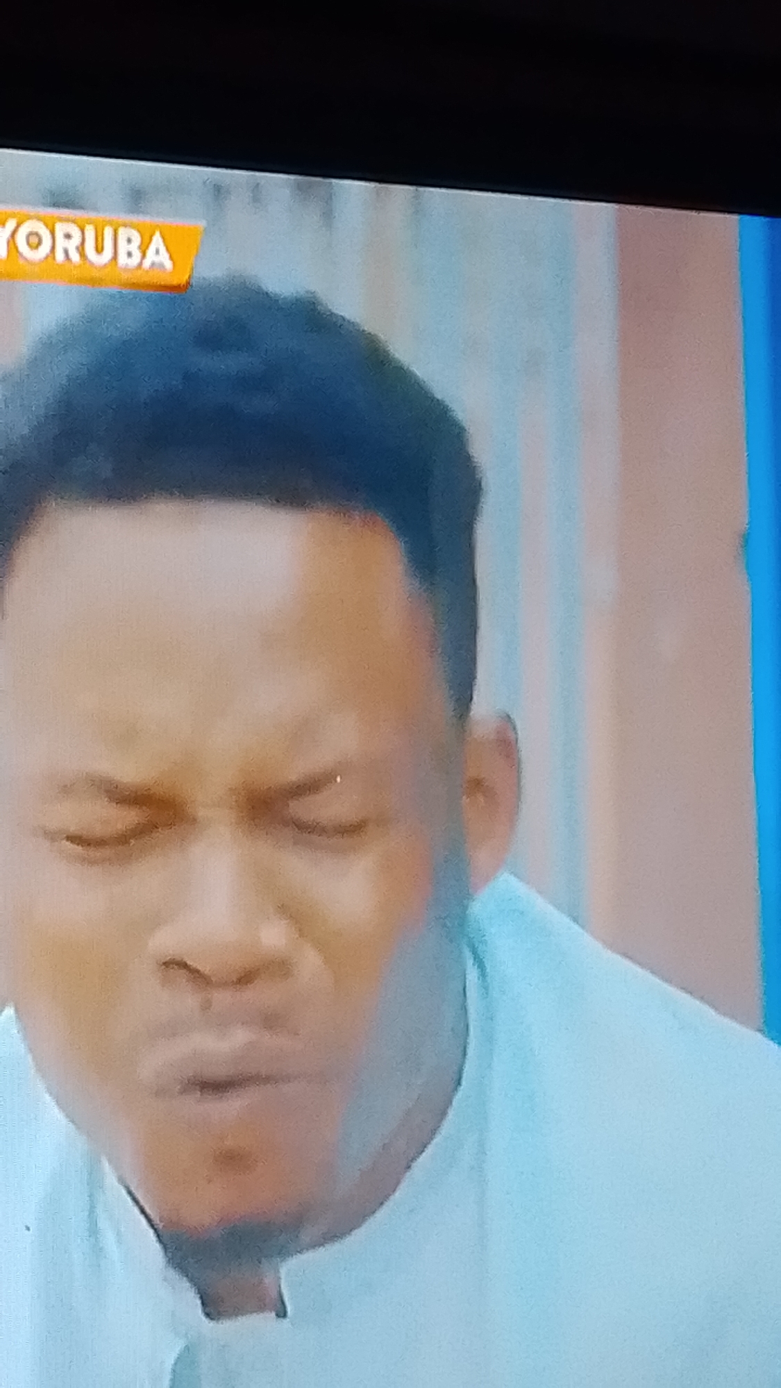 The image shows a close-up of a man's face. He is wearing a light blue collared shirt. His eyes are closed and his mouth is slightly open, as if he is speaking or reacting to something. Above him, an orange banner with the word "YORUBA" is visible. The background appears to be a wall with vertical stripes of light brown and white. The scene is likely from Lokoja, Nigeria, as indicated by the text on the banner, which could be a sign for a television program, a cultural event, or a place of business related to Yoruba culture.