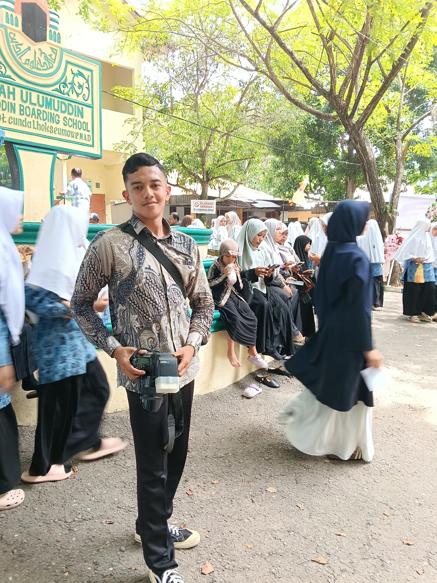 A young man stands holding a camera in front of the "AH ULUMUDDIN BOARDING SCHOOL" in Lhokseumawe, Indonesia. He is wearing a patterned shirt and dark pants. Behind him, a group of women, many wearing hijabs, are seated on a bench. One woman in a dark hijab and white skirt is walking towards the right side of the frame. A sign that says "DILARANG MEROKOK" (No Smoking) is visible in the background. There are trees overhead, casting shadows on the ground. The ground is paved. Other people in similar attire are visible in the background.