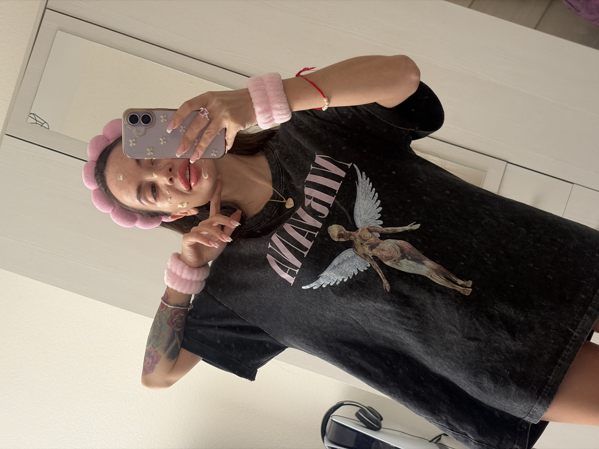 A young woman is captured in a mirror selfie, posing playfully indoors, likely in Saint Petersburg, Russia. She is wearing a dark, faded or acid-washed oversized t-shirt featuring a light pink, stylized "NIRVANA" text alongside an anatomical angel graphic with white wings, reminiscent of the band's "In Utero" album art.
She has a light pink, fluffy petal-shaped headband on her head and matching wristbands. Her face has dollops of a light-colored skincare product on her cheeks and chin, suggesting she is in the middle of a beauty routine. She is making a playful expression with her tongue slightly out and a peace sign gesture with her left hand. Her nails are long and manicured with a French tip style.
She holds a smartphone with a light lavender-colored case adorned with small white floral patterns. A ring with a large pink stone is visible on her right hand. Around her neck, she wears a delicate gold chain with a heart-shaped pendant. Her right wrist features a red string bracelet with white beads, while her left arm displays a colorful tattoo, primarily featuring pink floral designs, possibly roses, and a silver bracelet.
The background, visible as a mirror reflection, shows a simple, light-colored room interior, likely a bedroom or bathroom. In the bottom right corner, a pair of black and white headphones and what appears to be a gaming console (possibly a PlayStation 5) are visible on a white surface. The lighting is bright and even, indicating an indoor setting.