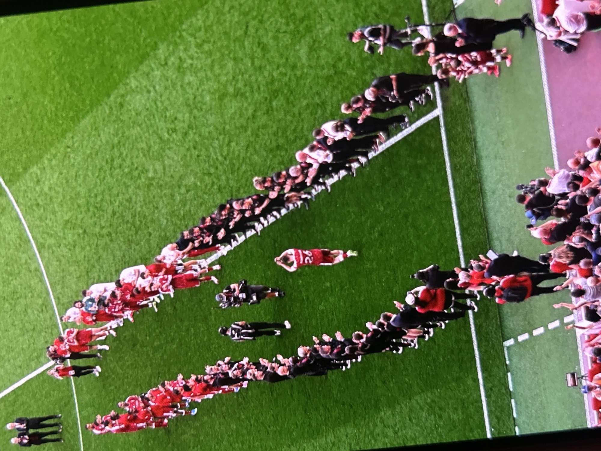 The image captures an aerial view of a football pitch, likely during a post-match event or ceremony. The main subjects are numerous people arranged in two long, curved lines on the green grass.
One line, predominantly composed of individuals in red and white football kits, stretches from the lower-left to the upper-middle of the frame. The other line, consisting of people in black suits or tracksuits, mirrors this curve, extending from the lower-right to the upper-middle. These lines appear to form a pathway or an honor guard.
In the center of the field, within the area created by these two lines, a single person wearing a red football jersey with the number "22" on the back is lying face down on the grass, seemingly in distress or exhaustion. Two other individuals in black attire are standing near this person.
Along the lines, some people are seen clapping, while others have their hands clasped or are standing solemnly. On the far right, beyond the white boundary lines of the pitch, a crowd of spectators is visible, and some individuals appear to be holding cameras or recording devices. Additional camera operators are present on the field near the lines of people.
The setting is an outdoor stadium under natural daylight. The grass is well-maintained with clear white markings. No specific weather conditions are discernible. The overall atmosphere appears to be a mix of ceremonial and emotional, possibly following a significant sporting event. The provided location context for this scene is Bania, Bulgaria.