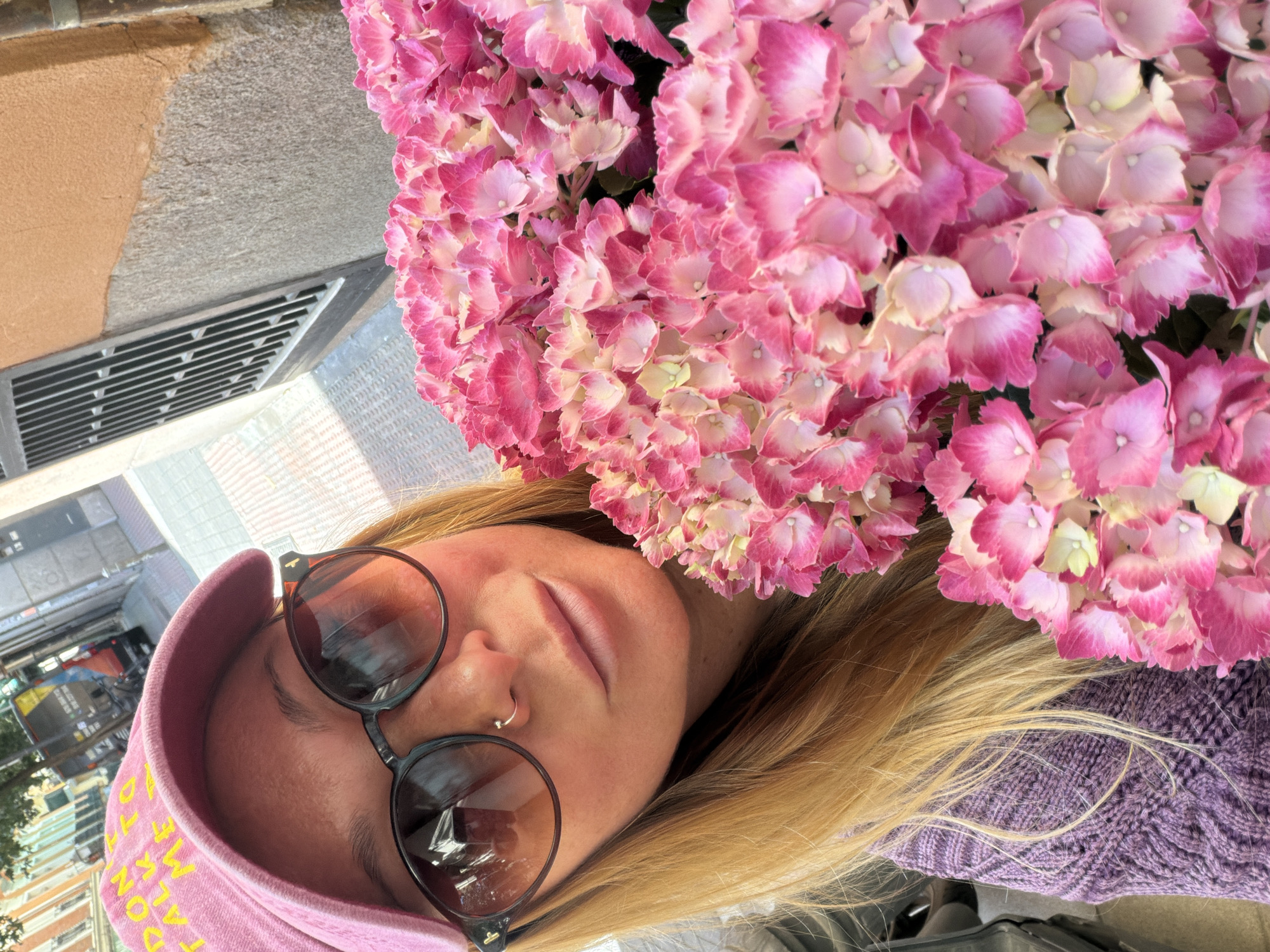 A rotated, close-up shot features a person with blonde hair and a pink hat adorned with writing. They are wearing sunglasses and have a nose ring. The person is positioned near a bouquet of pink hydrangeas. The background shows a building facade with a metal ventilation grill and street details, possibly indicative of a city environment. Given the urban context, the photo could plausibly be taken in Madrid, Spain, although there is no specific evidence to support this.