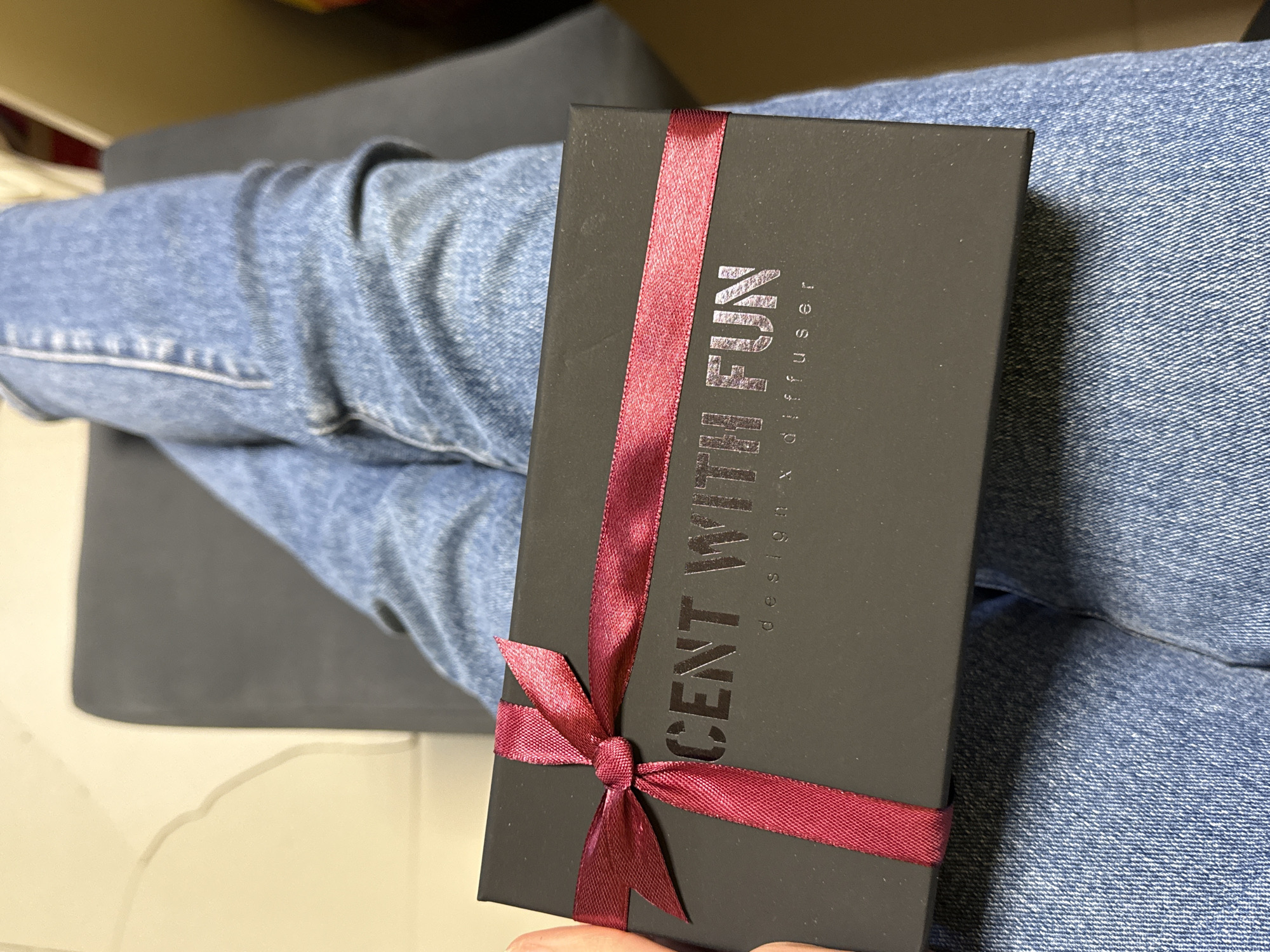 The image shows a black gift box with a burgundy ribbon, resting on the lap of a person wearing blue jeans. The box has text printed on it in grey. The setting appears to be indoors, with a grey cushion partially visible in the background.