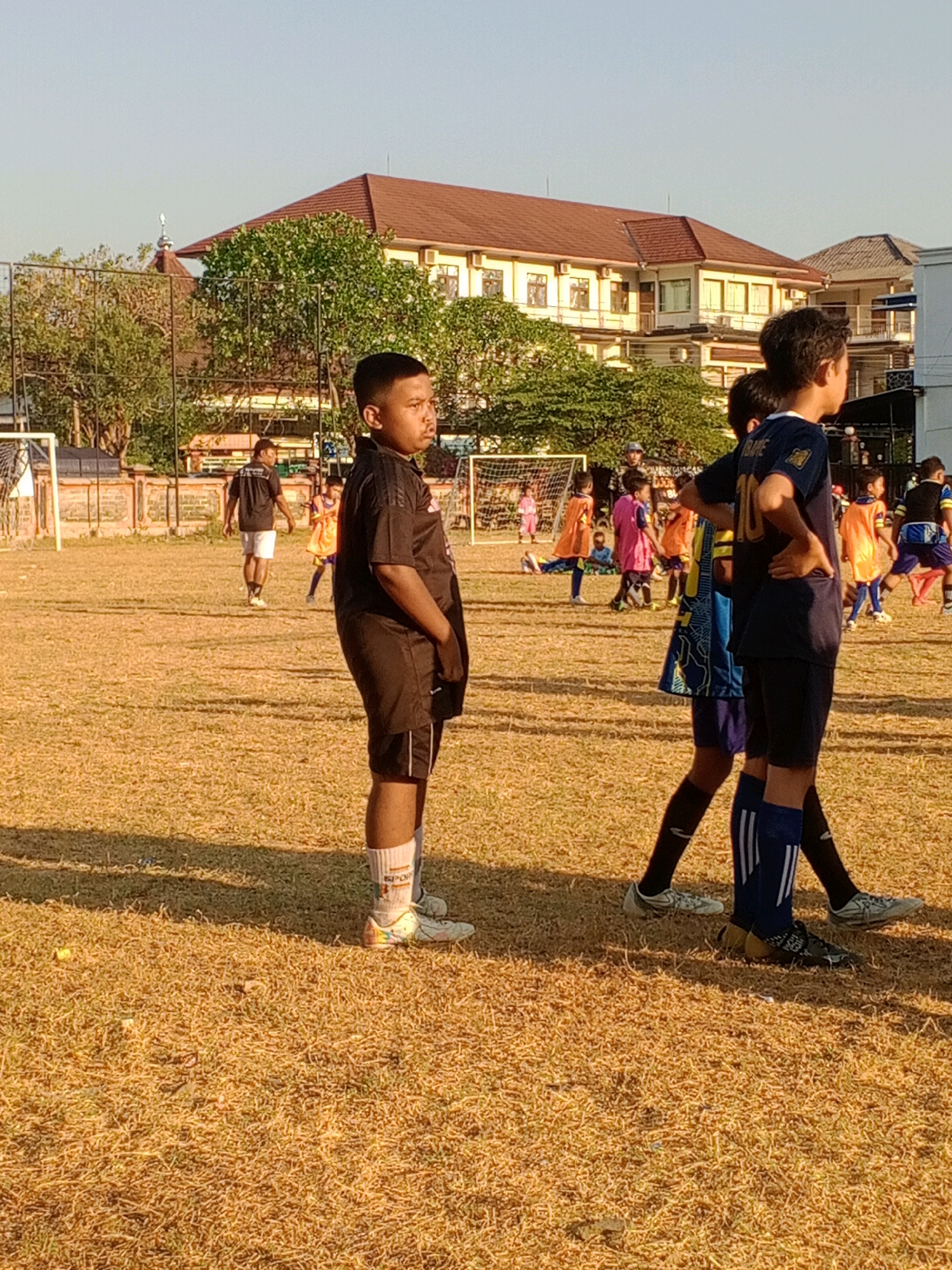 The image depicts an outdoor scene on a dry, grassy field, likely a sports ground, under a clear sky. The location is identified as Giritirto, Indonesia. In the foreground, a young boy wearing a black soccer jersey and shorts stands facing right, with his hands on his hips. He is wearing white socks with colorful accents and white soccer cleats. Behind him, other children, presumably part of a soccer team, are scattered across the field, some in orange and blue jerseys, some in white. In the middle ground, a soccer goal can be seen on the left, and a fence runs along the edge of the field. In the background, a large building with a red-tiled roof and cream-colored walls stands prominently, possibly a school or sports facility. Trees with lush green foliage are also visible around the building. The overall impression is that of a soccer practice or game taking place on a sunny day in an urban or suburban setting in Giritirto, Indonesia.