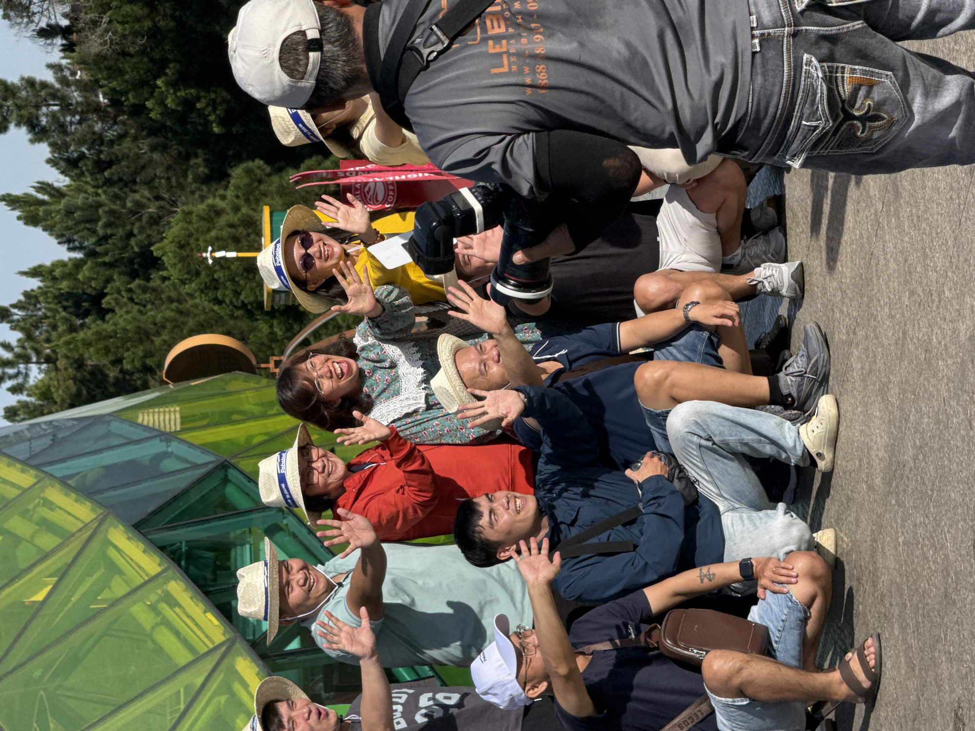 A group of people are posing for a photograph in front of a large, faceted, green and yellow glass structure, likely part of a tourist attraction. Most of the people are wearing straw hats. Several individuals are waving. One person is holding a professional camera with a flash, appearing to be the photographer. The background consists of lush green trees and a bright sky. The ground appears to be paved.
The scene strongly resembles the Tuyen Lam Lake area or a similar botanical garden or park setting in Da Lat, Vietnam, given the architectural style of the structure and the presence of coniferous trees common in the region. The overall atmosphere is casual and festive.