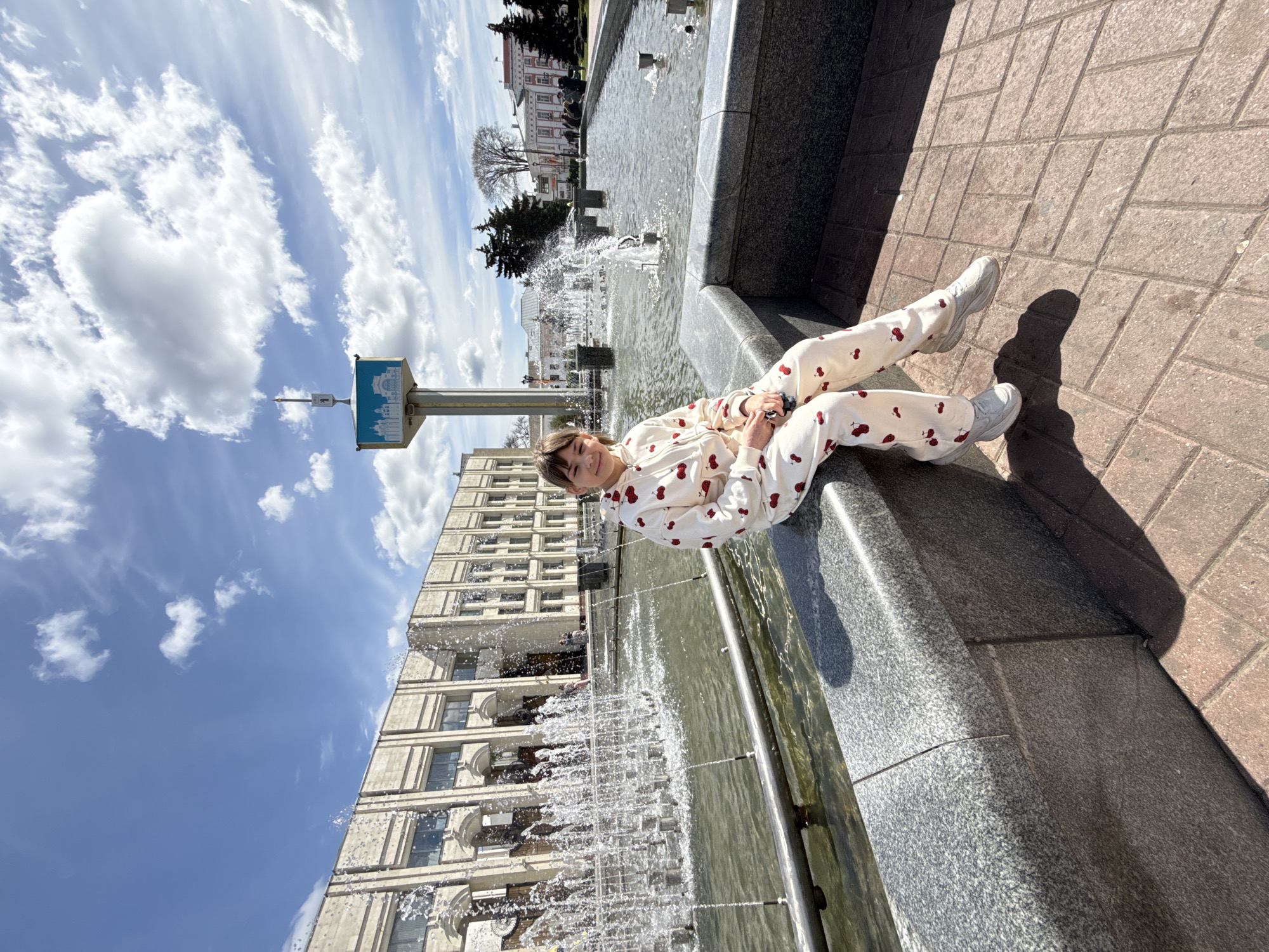A young woman is lying on the edge of a fountain, wearing a cherry-patterned tracksuit and white sneakers. She is smiling and facing the camera. The fountain is located in a city, with a tall building behind it and a monument to the left. The weather appears sunny with some clouds in the sky. The surrounding area is paved with brick.