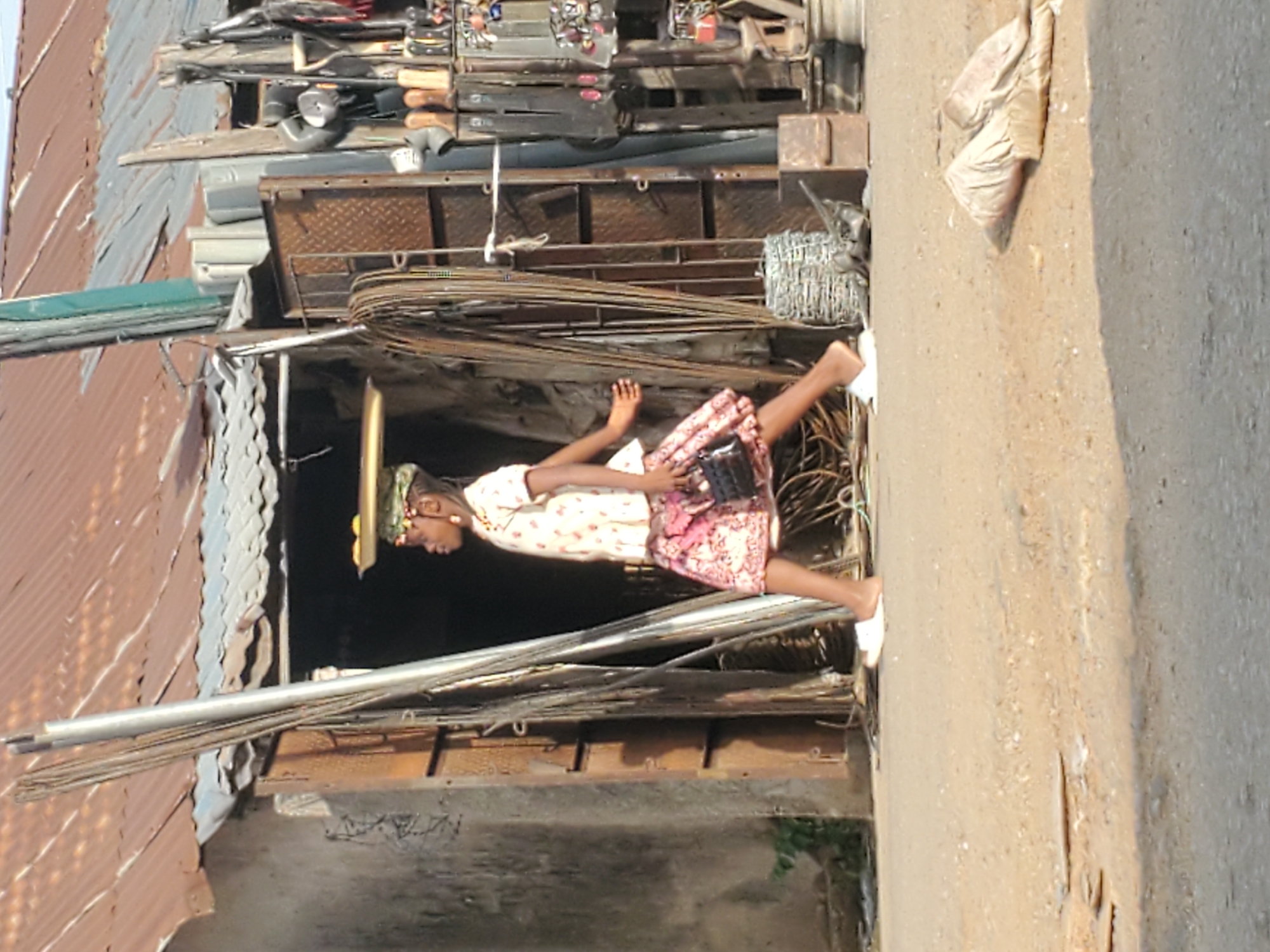 In Mararaba, Nigeria, a young girl in a light-colored patterned dress, barefoot, is standing in the doorway of a corrugated iron structure cluttered with metal materials. She holds a dark object in her hands. The structure is located alongside a concrete road. Metal items are stored above the doorway.