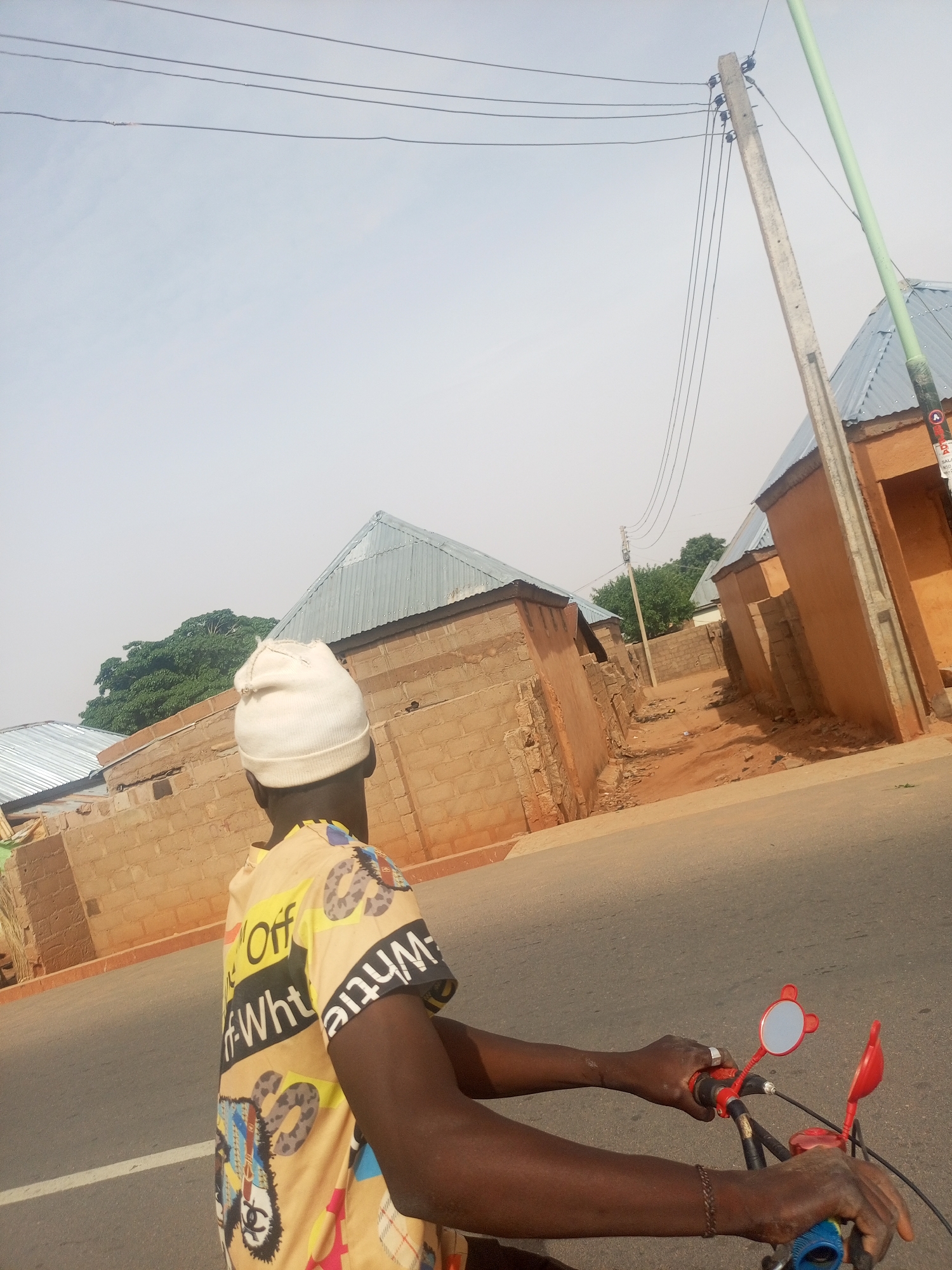A man wearing a white beanie and a yellow and black patterned shirt is riding a bicycle down a paved road in what appears to be a residential area. The man's dark skin contrasts with the light-colored beanie and shirt. He is holding the handlebars of the bicycle, which has red-framed mirrors. In the background are brick buildings with corrugated metal roofs. A utility pole with electrical wires is visible on the right side of the image. The sky is pale blue and overcast. The location is Tempure, Nigeria.