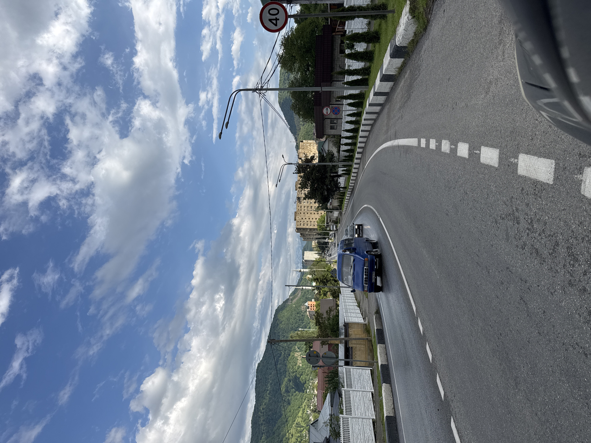 A blue minibus drives down a wet asphalt road in Karachayevsk, Russia. The road is bordered by a white fence with black and white striped concrete barriers on the right. In the background, green mountains rise behind a town with buildings, including a mosque with minarets. The sky is a bright blue with scattered white clouds. A speed limit sign indicating 40 is visible on the right side of the road.