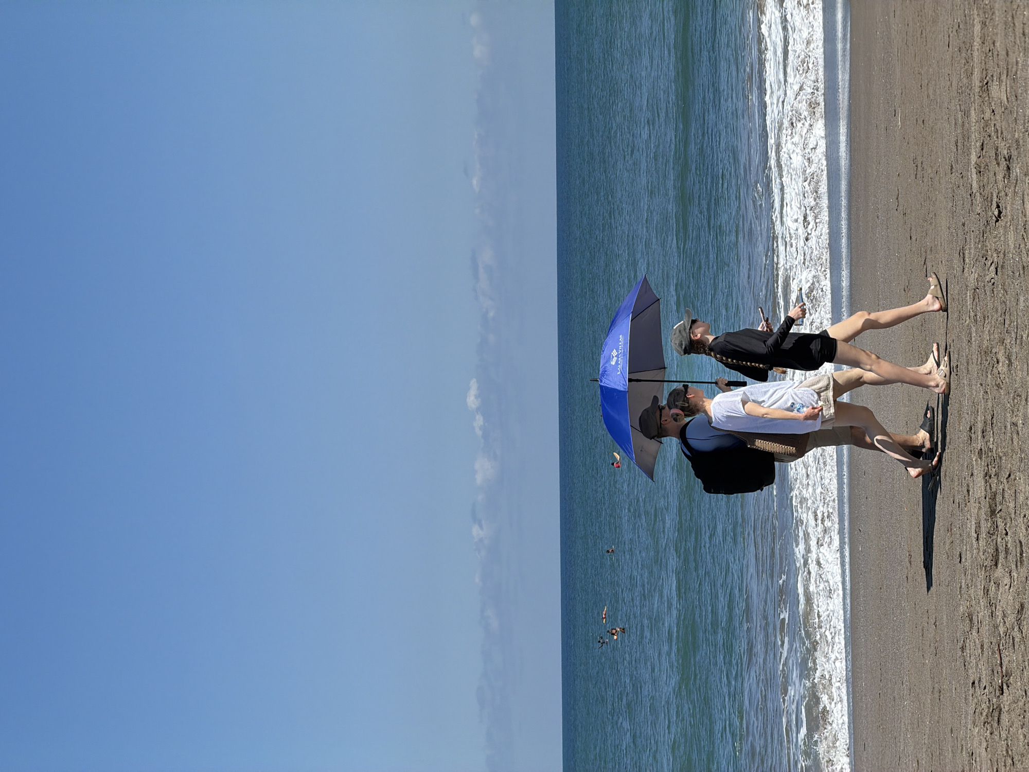 The image captures a beach scene, likely in North Kuta, Indonesia, where the sky, ocean, and beach are distinctly divided into segments. Two individuals are standing on the sandy shore. One is holding a blue umbrella, while the other is looking at their phone. The ocean water appears to be lapping onto the beach. Other people can be seen swimming in the ocean.
