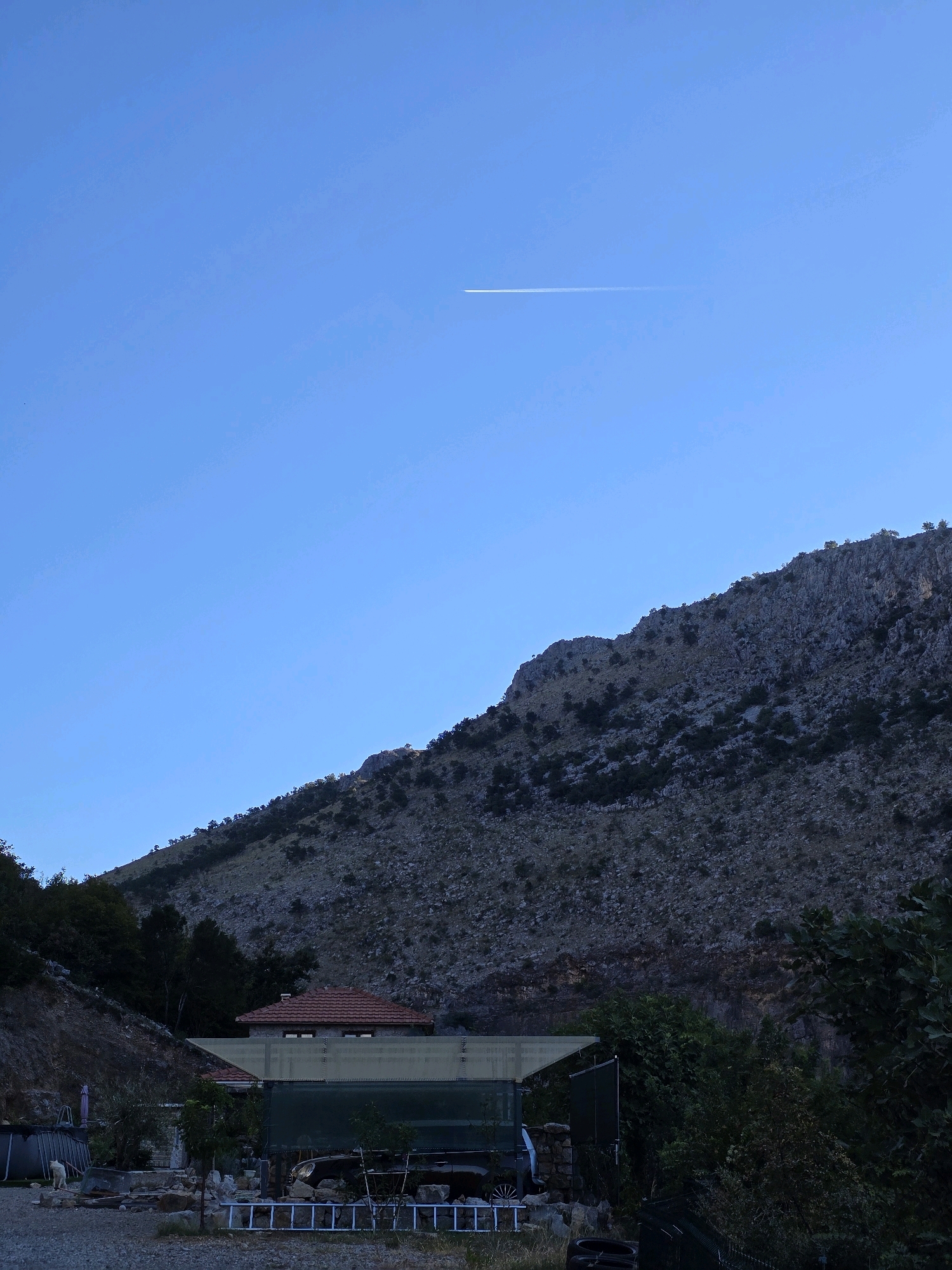 An image captures a clear, blue sky with a faint contrail from an airplane high above. The sky dominates the upper portion of the frame, stretching over a rugged, rocky mountain slope. The mountain is sparsely covered with dark green trees and vegetation, giving it a textured, uneven appearance. In the foreground, a residential structure with a red-tiled roof is partially visible, nestled at the base of the mountain. The building appears to be a house or a small dwelling. Various objects are scattered around the immediate vicinity of the house, including what looks like an inflatable pool on the left, some gardening tools or equipment, and a parked vehicle. The ground is a mixture of gravel and dirt. The overall scene suggests a rural or semi-rural setting, possibly on the outskirts of a city like Podgorica, Montenegro, where mountainous terrain meets residential areas. The lighting indicates it might be late afternoon or early evening, given the gentle illumination and the soft shadows. There are no visible people in the image.
