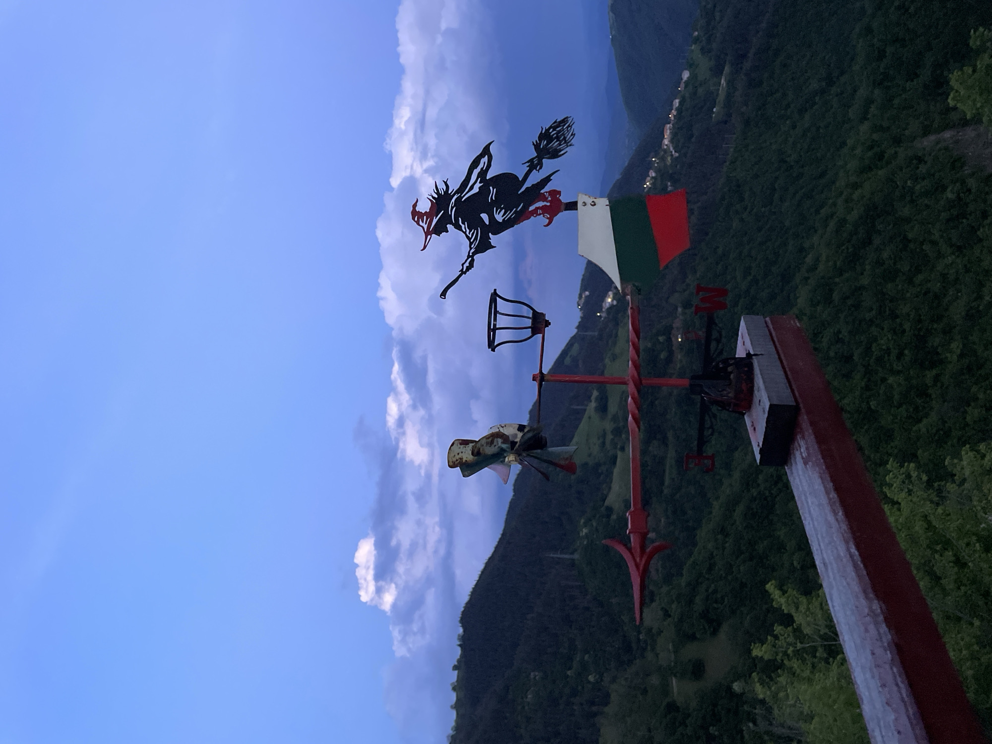 The image shows a weathervane mounted on a red wooden post in an outdoor setting, possibly in the vicinity of Pechinska, Bulgaria. The weathervane features a black silhouette of a witch riding a broomstick, and below the witch there is what appears to be a metal lantern. The landscape beyond includes a lush green mountainside with settlements visible in the background. The sky is a mix of clear blue and clouds.