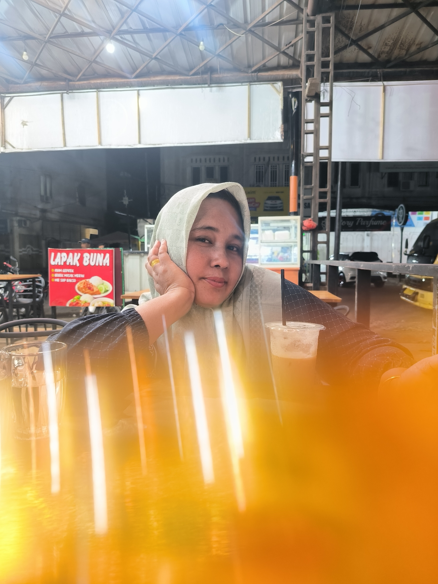 A woman wearing a light-colored hijab sits at an outdoor table with drinks, near a food stall called "LAPAK BUNA" in Lhokseumawe, Indonesia. The scene appears to be at night. A clear glass mug with a dark liquid is visible, along with a plastic cup of iced coffee. A ring is visible on her finger. The table and foreground are partially obscured by a bright, orange glare. In the background, there is a row of buildings and vehicles, including a food stall with visible signage advertising "BUNA GEPREK" and other dishes. There is an advertisement board on a wall. The area has tables and chairs.